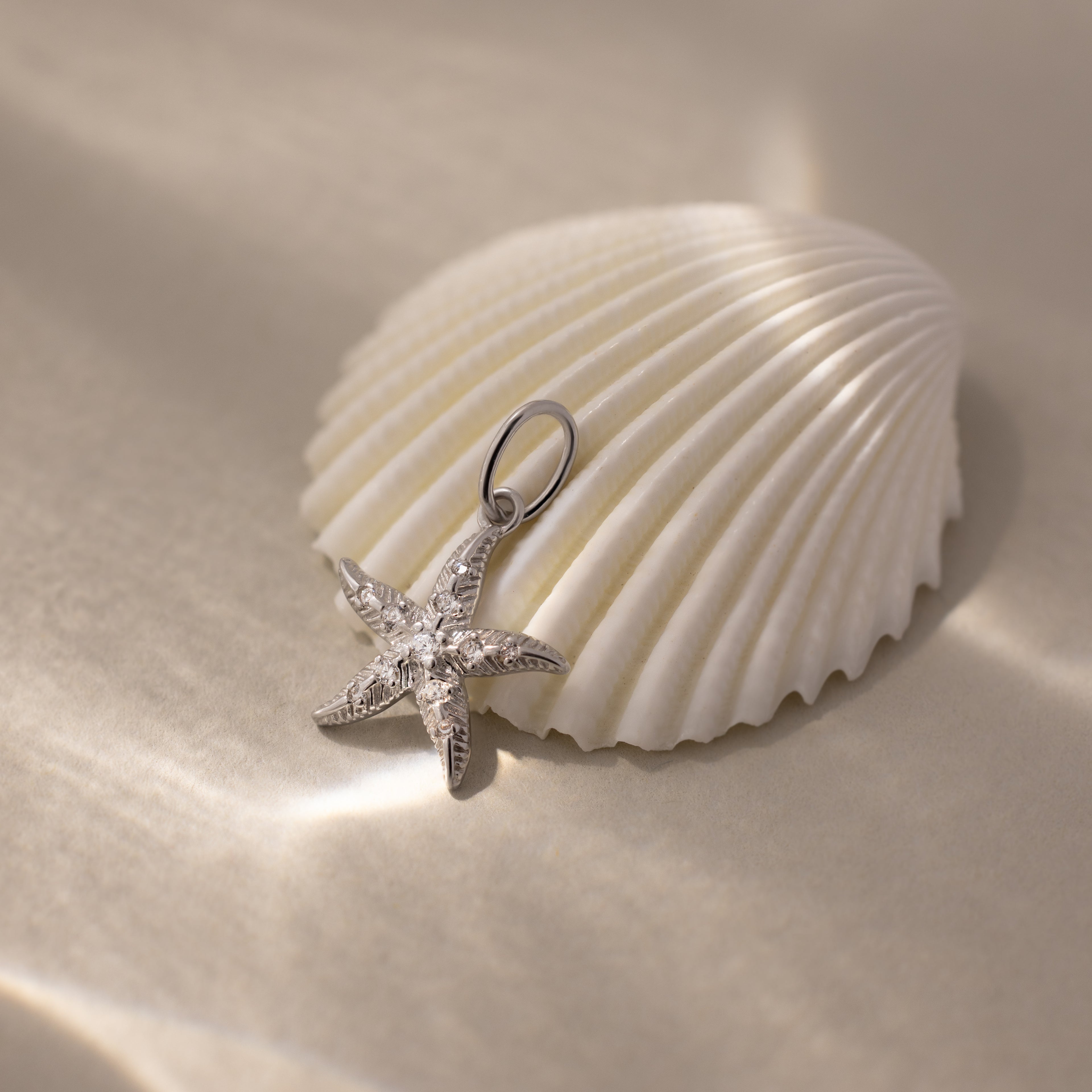 The Pave Starfish Charm shines atop a white seashell against a soft, shimmering background, capturing genuine coastal charm.
