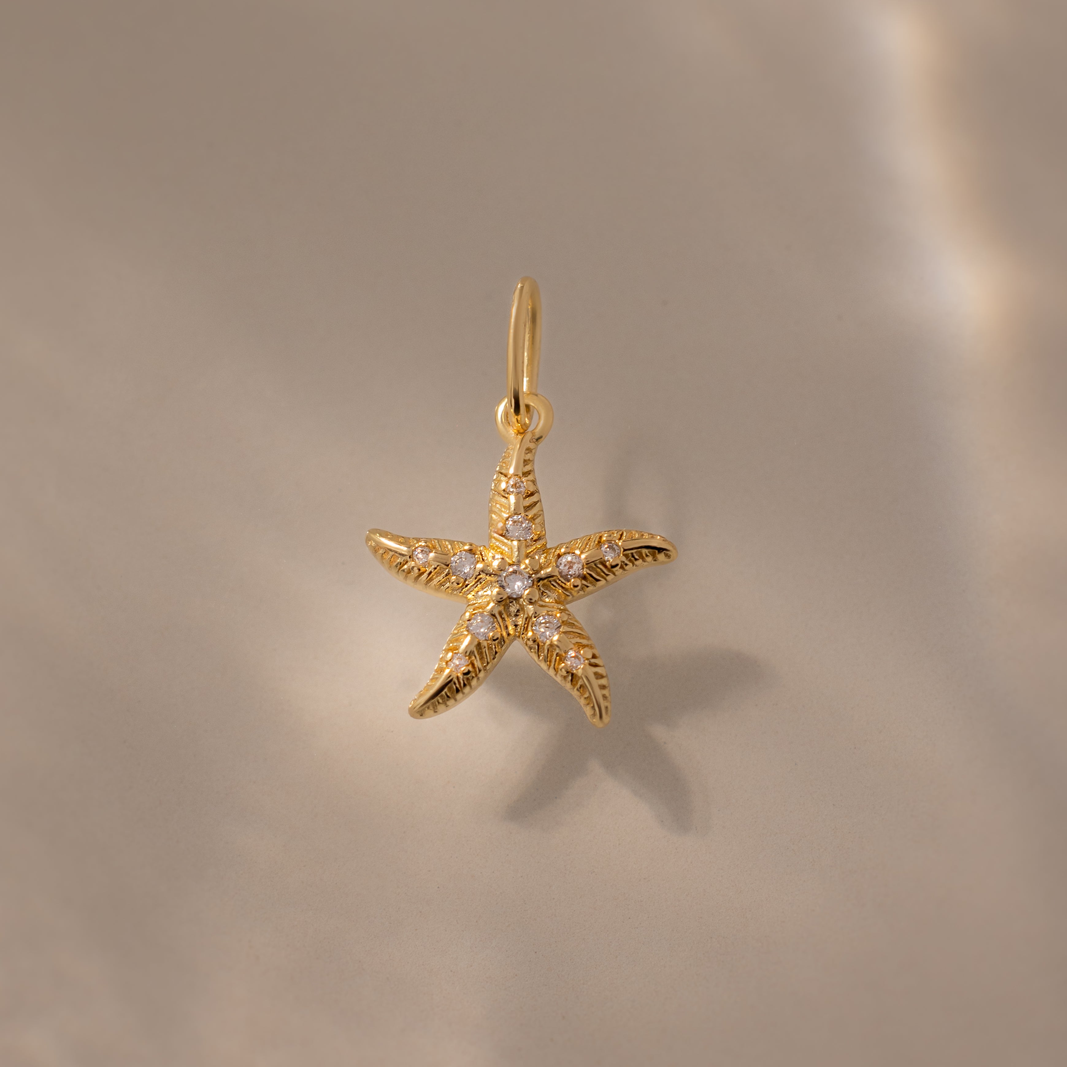 The Pave Starfish Charm in 18K Gold features shimmering gemstones, photographed on a beige surface with soft light—a stunning piece of coastal-inspired jewelry.