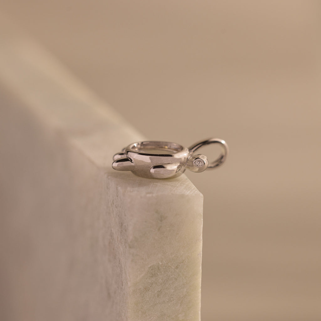 The Diamond Turtle Charm features a shimmering silver finish and small gemstone, resting atop light marble to embody the beauty of ocean-inspired jewelry.