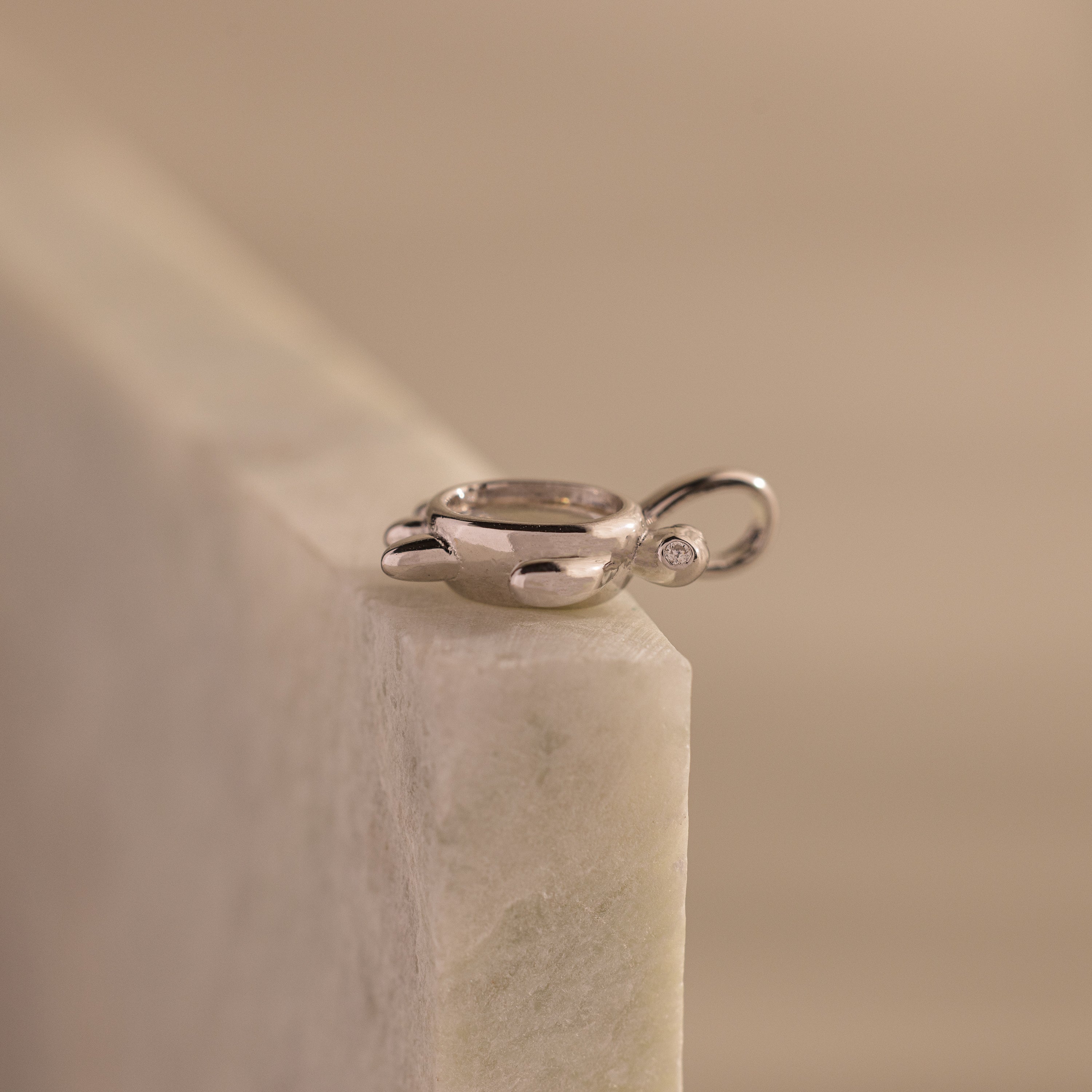 The Diamond Turtle Charm features a shimmering silver finish and small gemstone, resting atop light marble to embody the beauty of ocean-inspired jewelry.