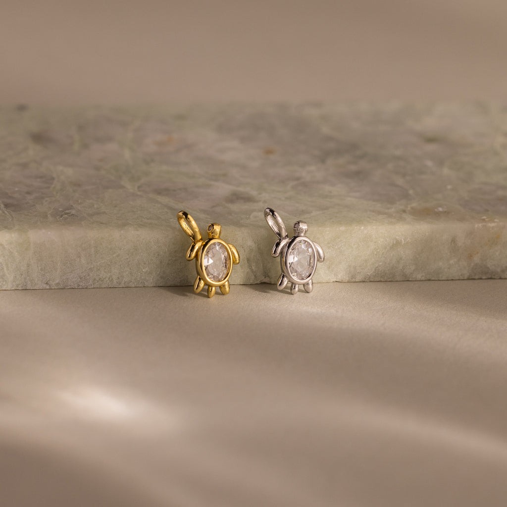 Two Diamond Turtle Charms—one gold, one silver—rest on a marble surface under soft lighting, each featuring a delicate turtle design ideal for ocean jewelry lovers.