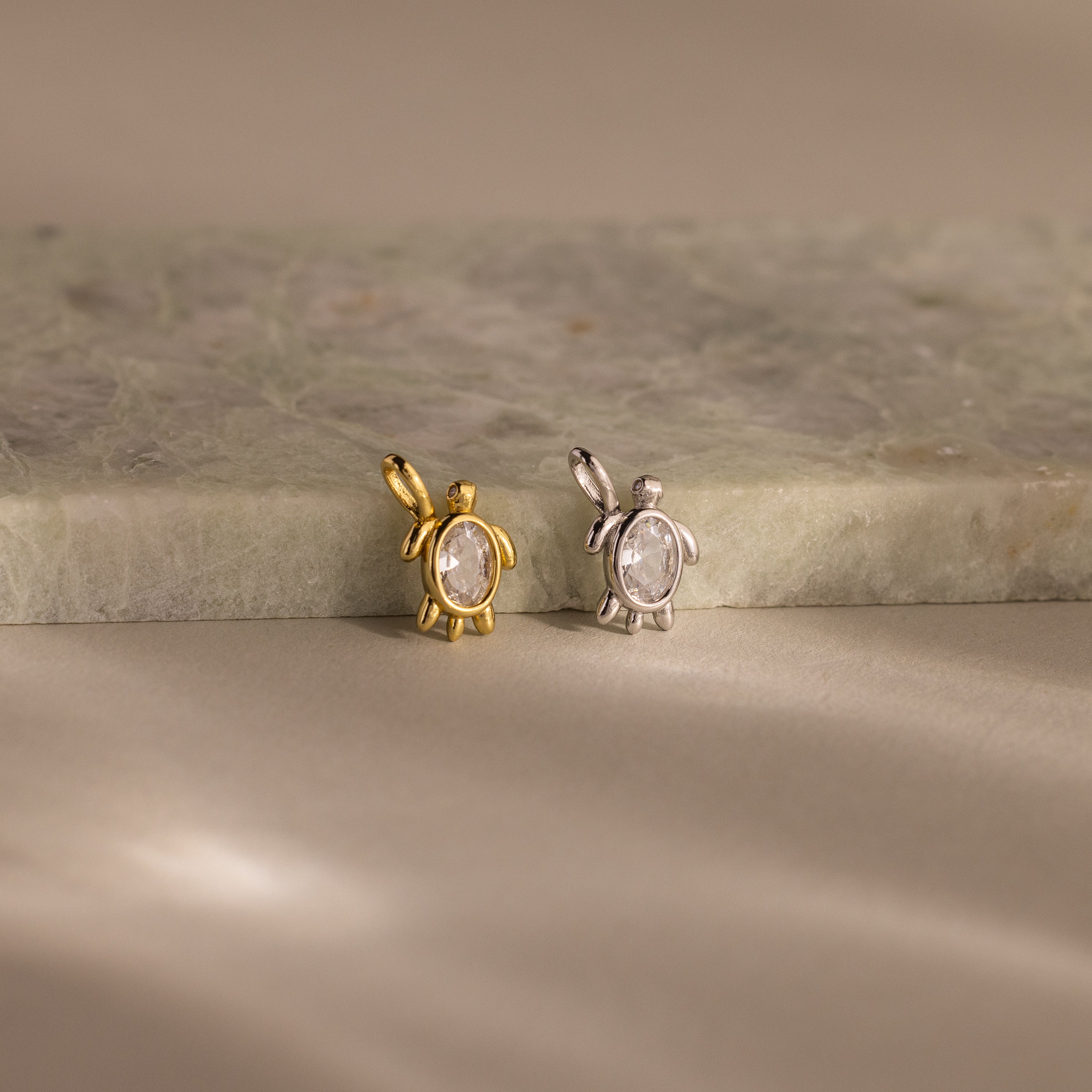 Two Diamond Turtle Charms—one gold, one silver—rest on a marble surface under soft lighting, each featuring a delicate turtle design ideal for ocean jewelry lovers.