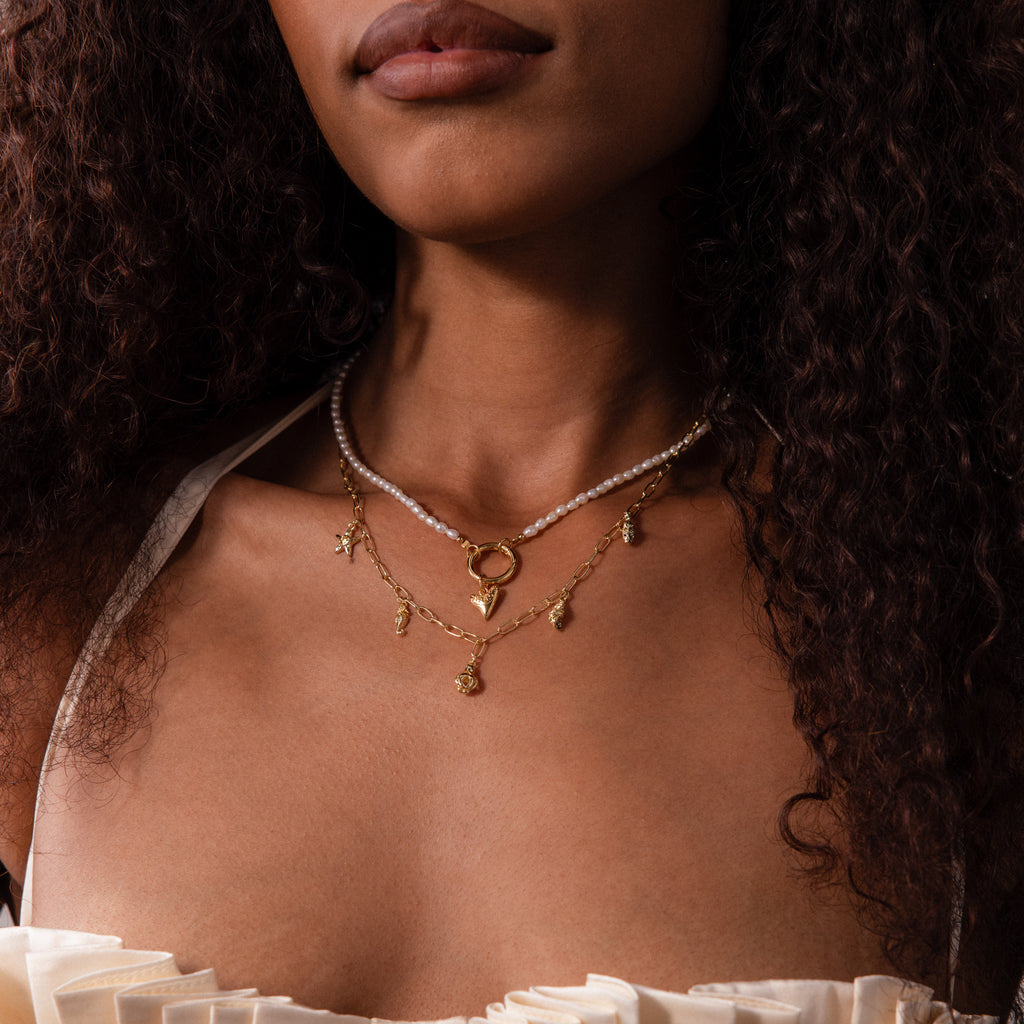 A woman with curly hair wears layered necklaces, including the Ocean Charm Necklace in 18K Gold and pearls, styled beachy and shown from shoulders to lips.