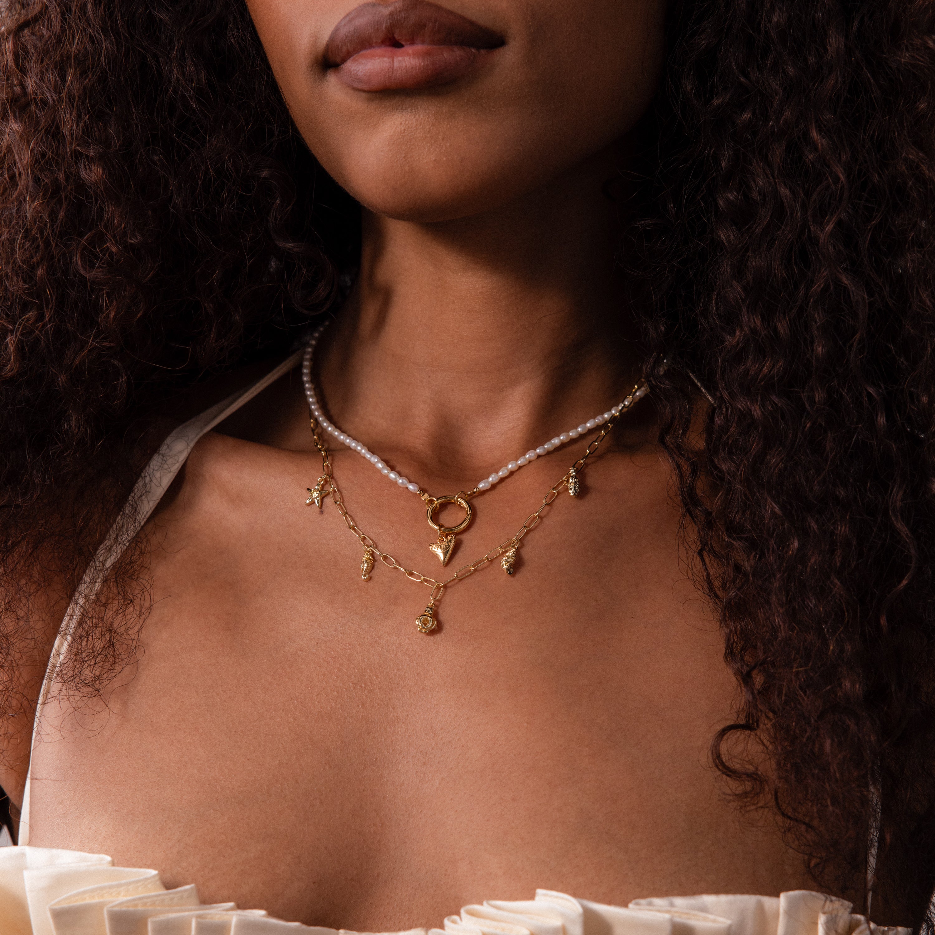 A woman with curly hair wears layered necklaces, including the Ocean Charm Necklace in 18K Gold and pearls, styled beachy and shown from shoulders to lips.