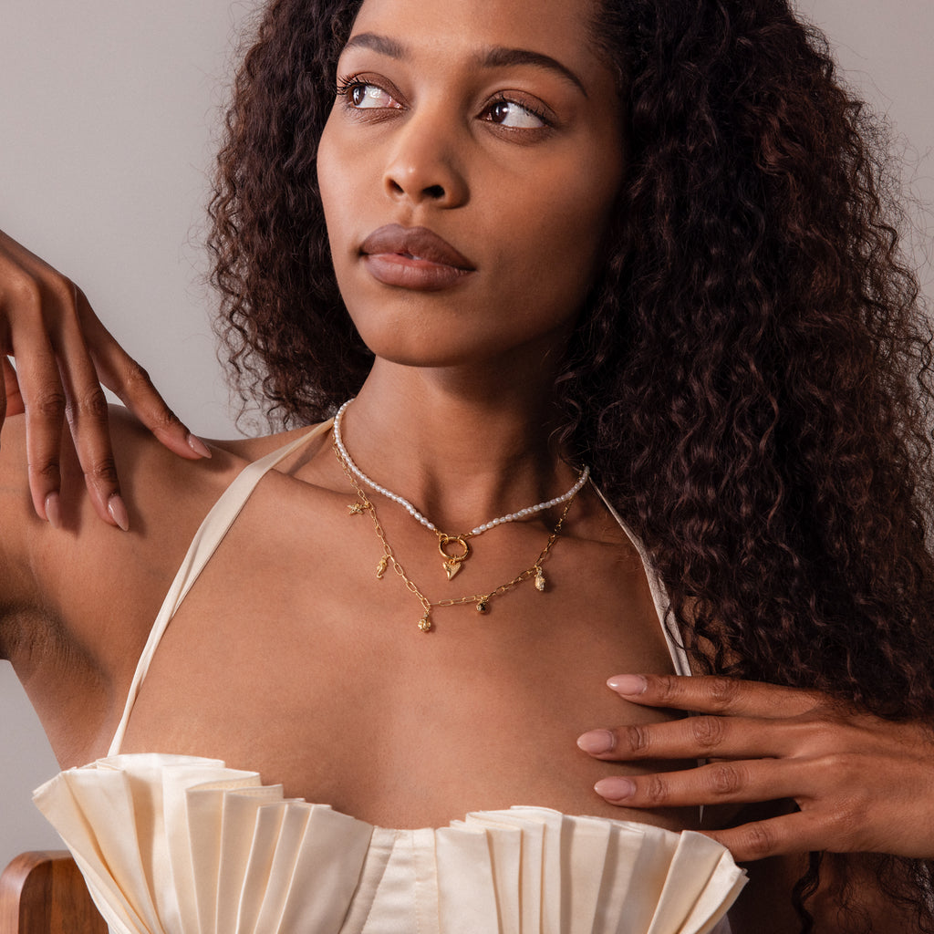 A woman with long curly hair wears layered gold necklaces, including the Ocean Charm Necklace in 18K Gold, paired with a cream pleated top as she gazes to the side.