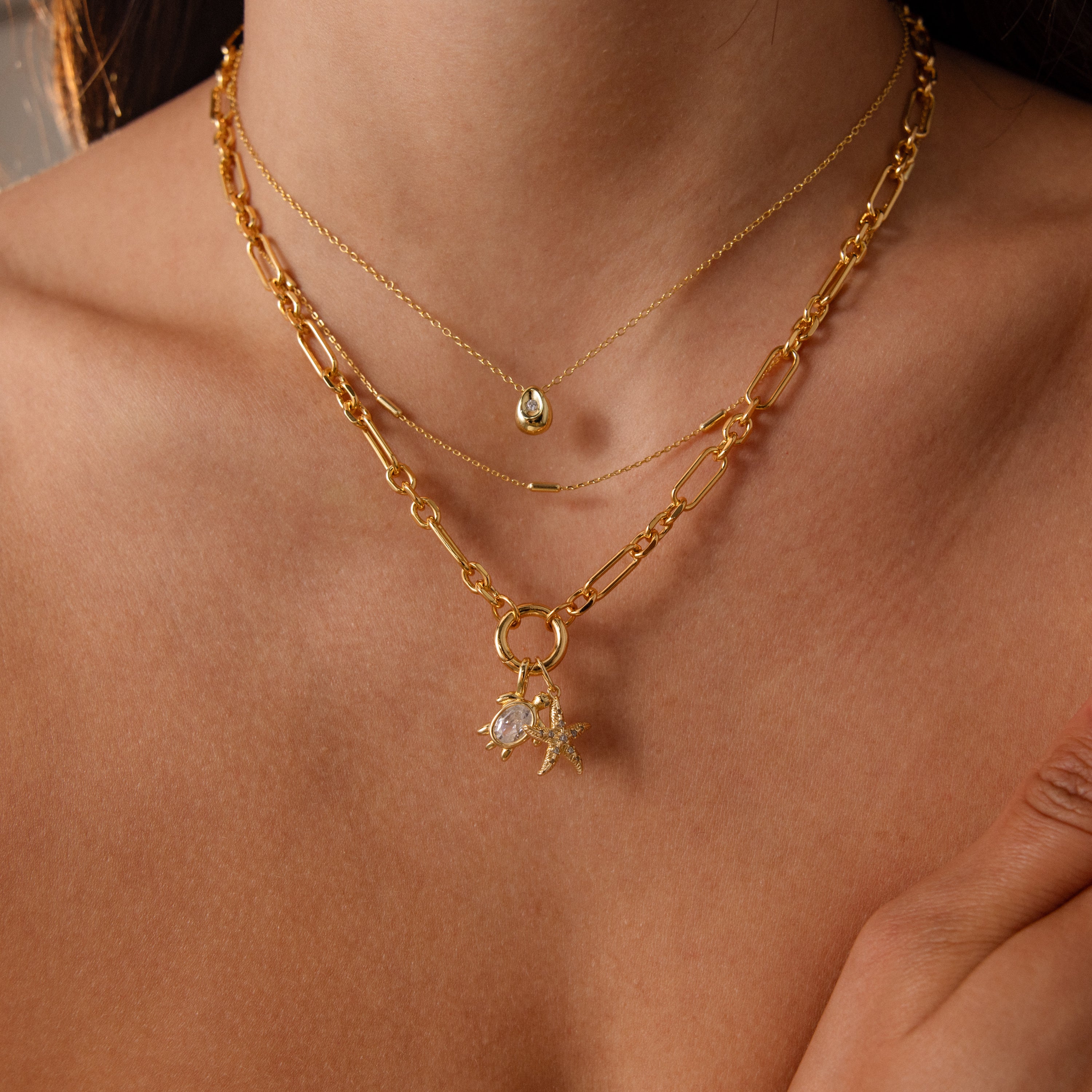 A woman wears layered gold necklaces, including the Diamond Turtle Charm in 18K Gold with a diamond-detailed shell and another necklace adorned with ocean-inspired charms and pearls.