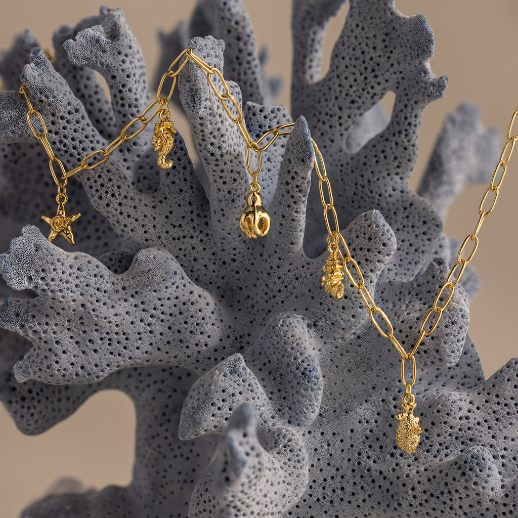 The Ocean Charm Necklace in 18K Gold, featuring sea-inspired pendants on textured grey coral, makes the perfect beachy necklace or summer jewelry accessory.