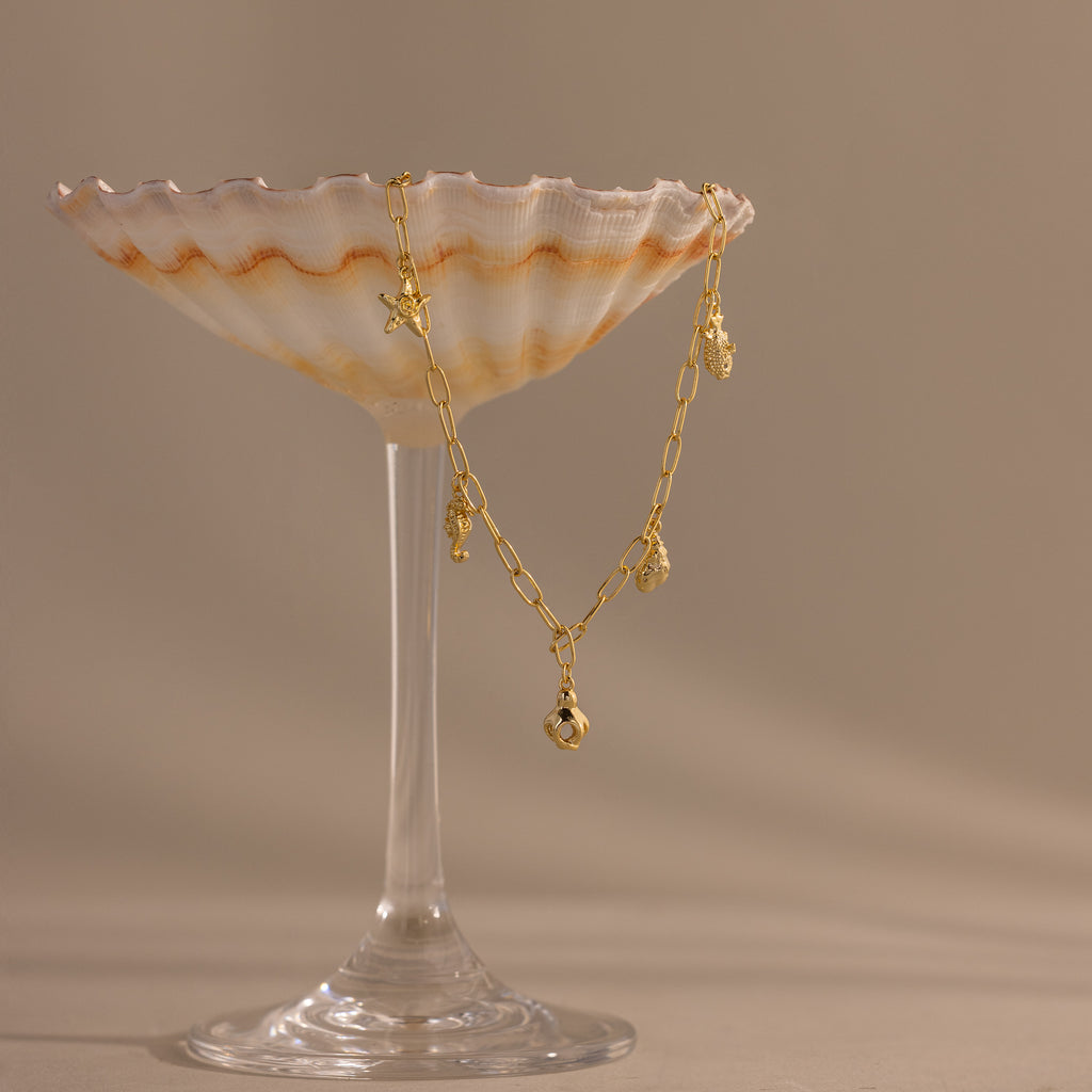 A gold charm bracelet draped over a seashell-shaped glass dish on a beige background, perfect for pairing with your favorite summer jewelry or the Ocean Charm Necklace in 18K Gold.