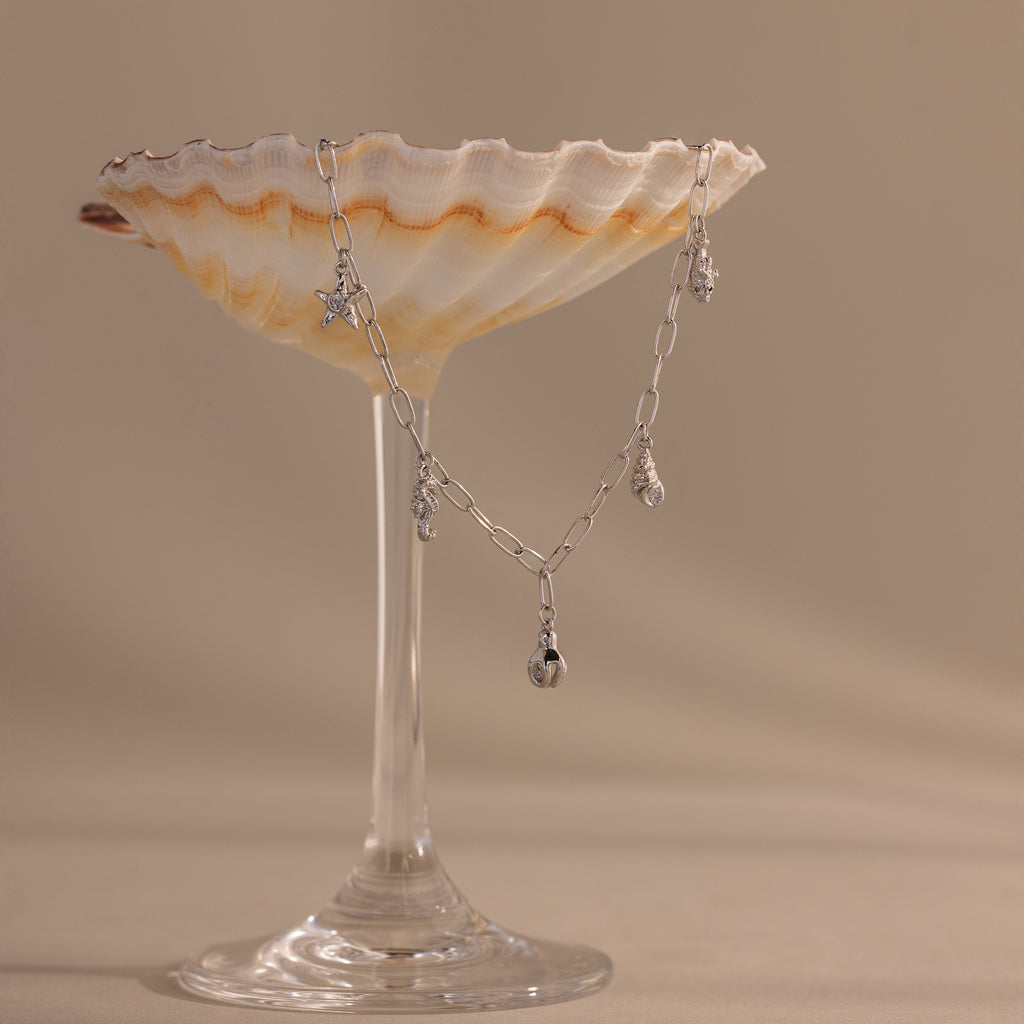 The Ocean Charm Necklace in Silver, displayed over a scallop shell on a glass stem against a beige backdrop—ideal for lovers of seaside jewelry.