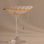 The Ocean Charm Necklace, in silver with seaside charms, is draped over a scallop shell and displayed on a glass stand against a beige background.