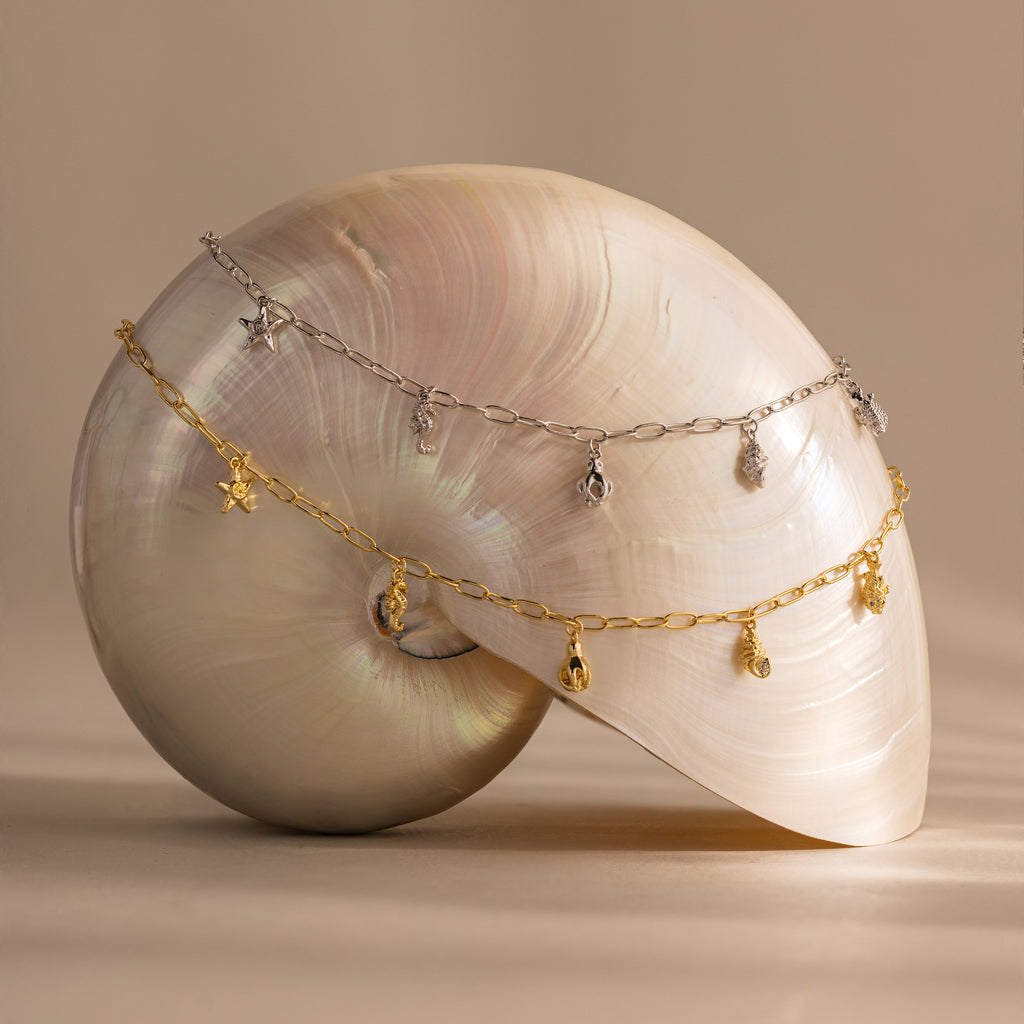 The Ocean Charm Necklace in Silver is displayed draped over a large iridescent seashell against a neutral background.