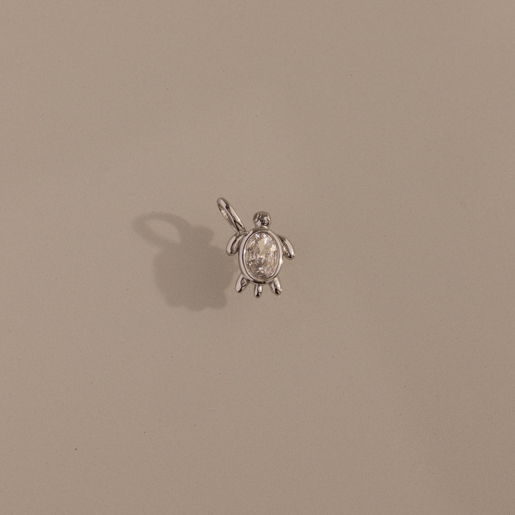 The Diamond Turtle Charm in Silver features a small turtle design with a loop, casting a shadow on beige—ideal for fans of ocean-inspired jewelry.