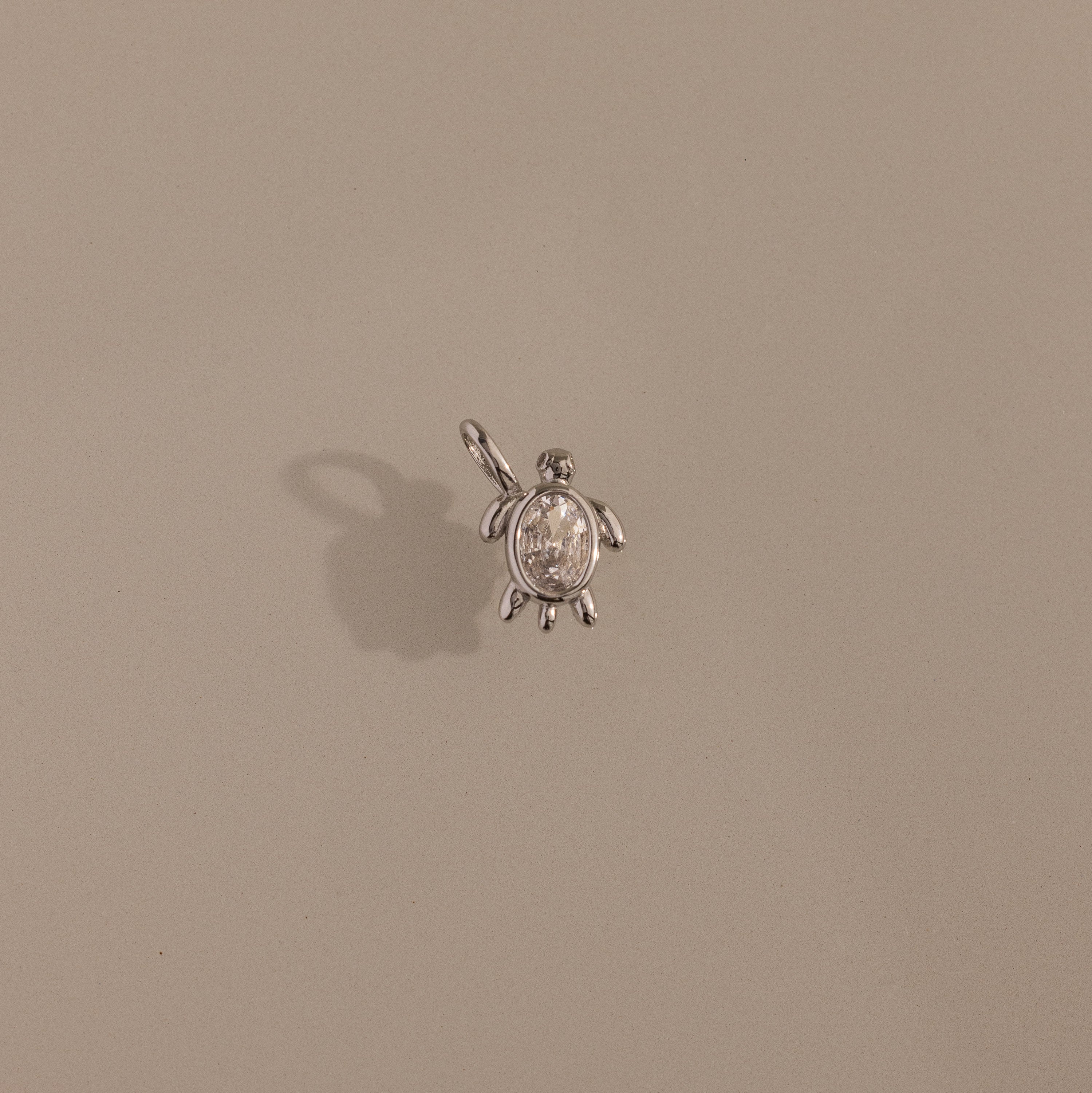 The Diamond Turtle Charm in Silver features a small turtle design with a loop, casting a shadow on beige—ideal for fans of ocean-inspired jewelry.