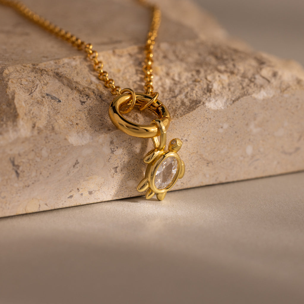 The Diamond Turtle Charm in 18K Gold features a diamond-detailed shell and rests elegantly on a textured stone, showcasing ocean-inspired jewelry at its finest.