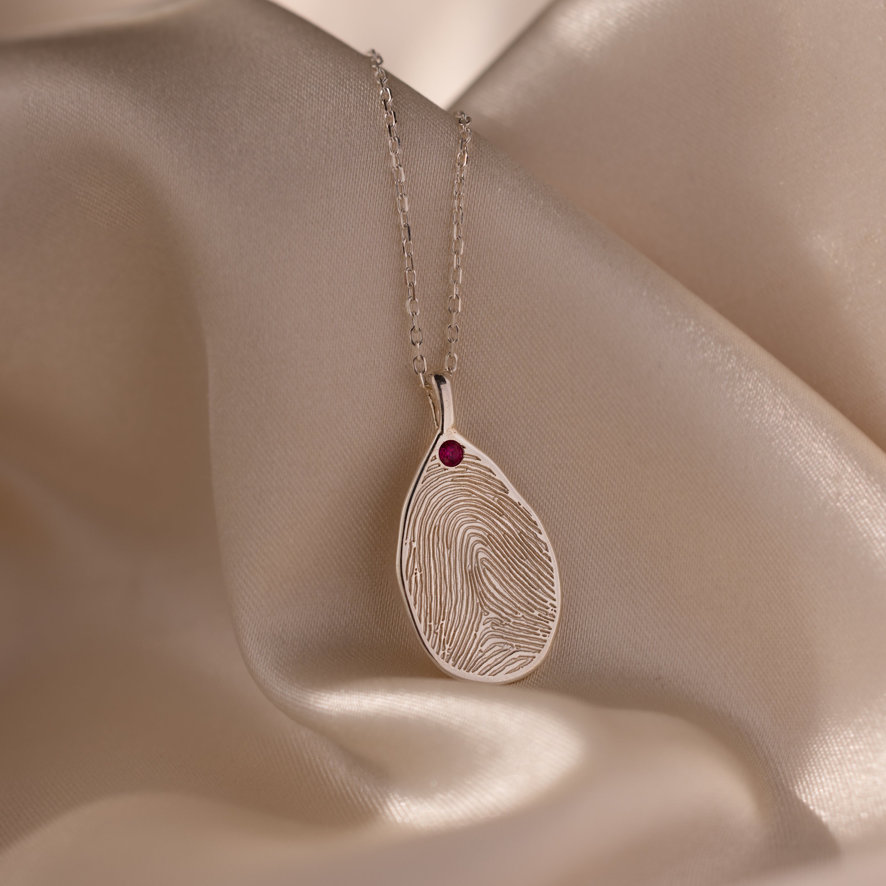 Fingerprint Birthstone Teardrop Necklace in Sterling Silver featuring a fingerprint pendant and a red gemstone, displayed on soft beige satin—a timeless keepsake, ideal for anyone who loves personalized jewelry.