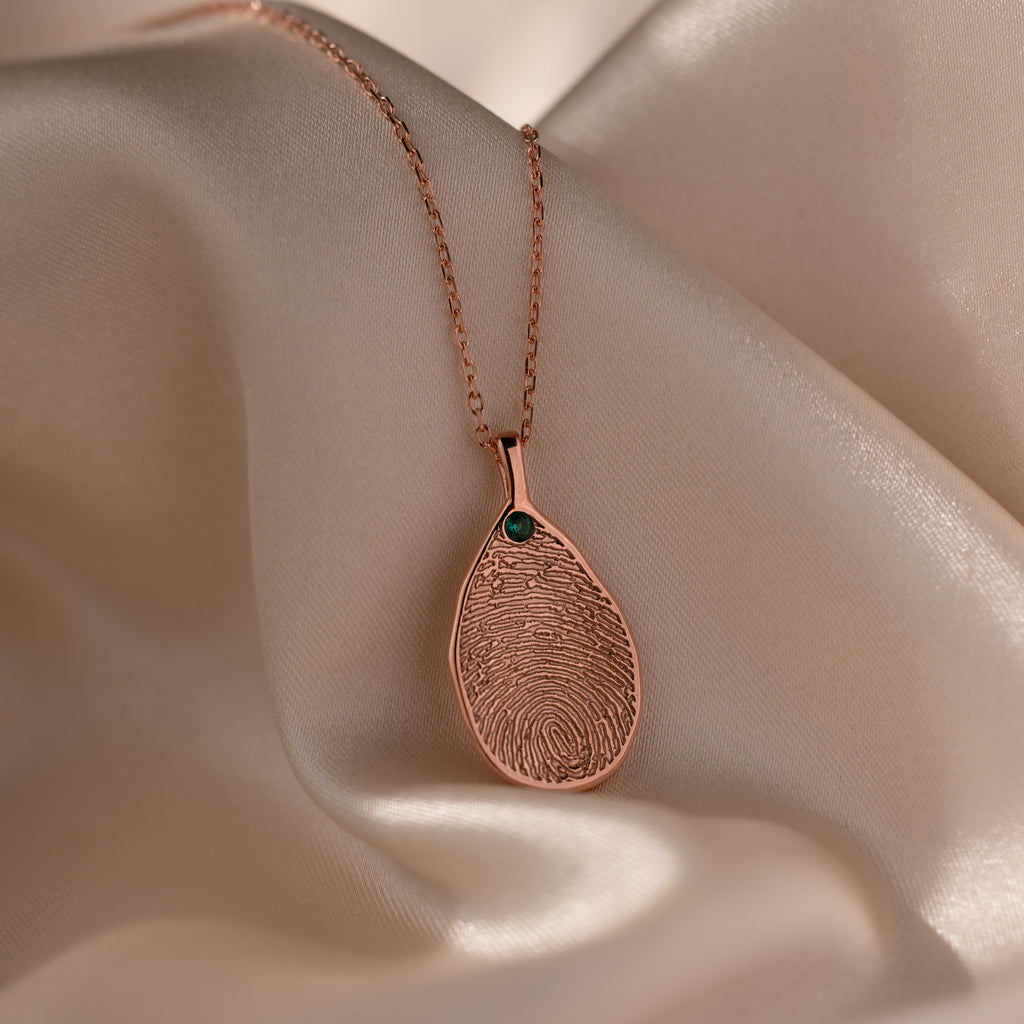 The Fingerprint Birthstone Teardrop Necklace in Rose Gold features a unique fingerprint design and small green gemstone on cream satin, making it an elegant personalized jewelry piece or meaningful gift.