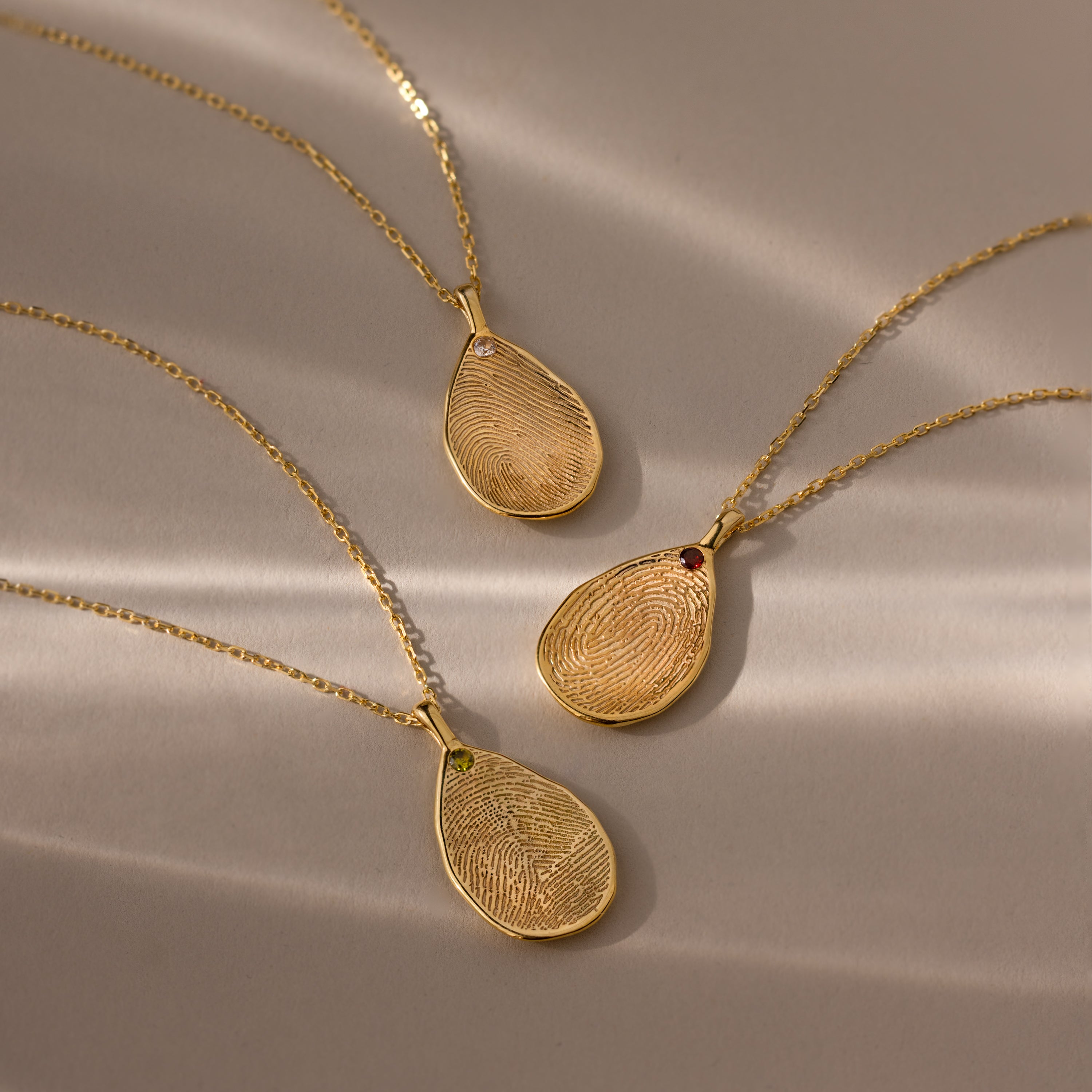 Three Fingerprint Birthstone Teardrop Necklaces in 18K Gold with custom fingerprint designs and gemstones are displayed on a beige background—each one a unique keepsake, ideal for those who love personalized jewelry.