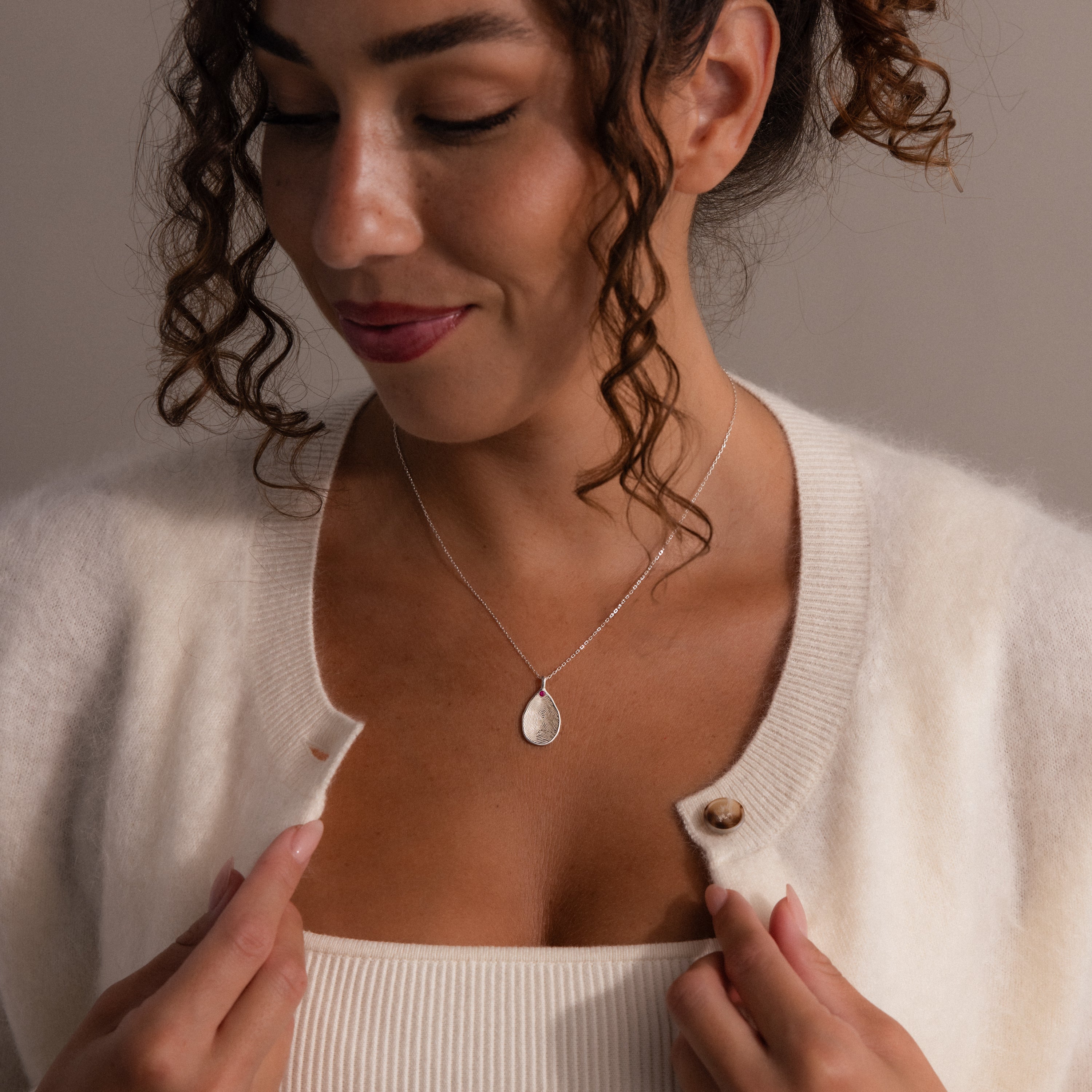 Woman smiling and looking down, wearing a white cardigan, a tank top, and the Fingerprint Birthstone Teardrop Necklace in Sterling Silver—a personalized jewelry piece that adds meaning to her look.
