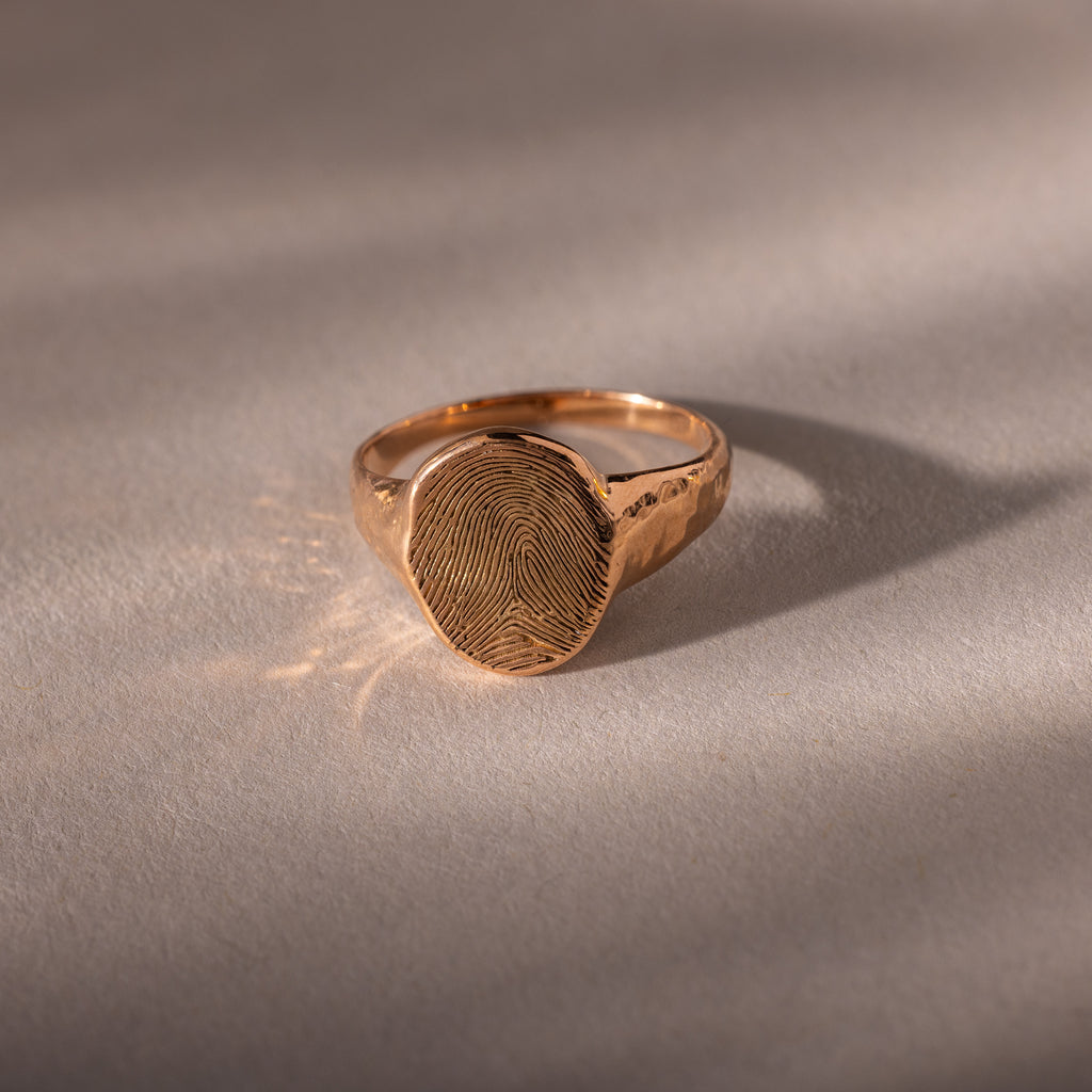 The Organic Fingerprint Signet Ring in Rose Gold features a round face engraved with a fingerprint, displayed on a soft, neutral surface—a distinctive piece of fingerprint jewelry.