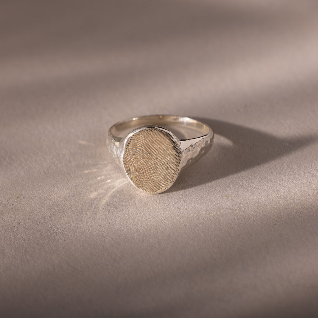 The Organic Fingerprint Signet Ring in Sterling Silver features a round bezel engraved with a fingerprint, creating a unique, personalized piece of jewelry that serves as a meaningful wearable keepsake.