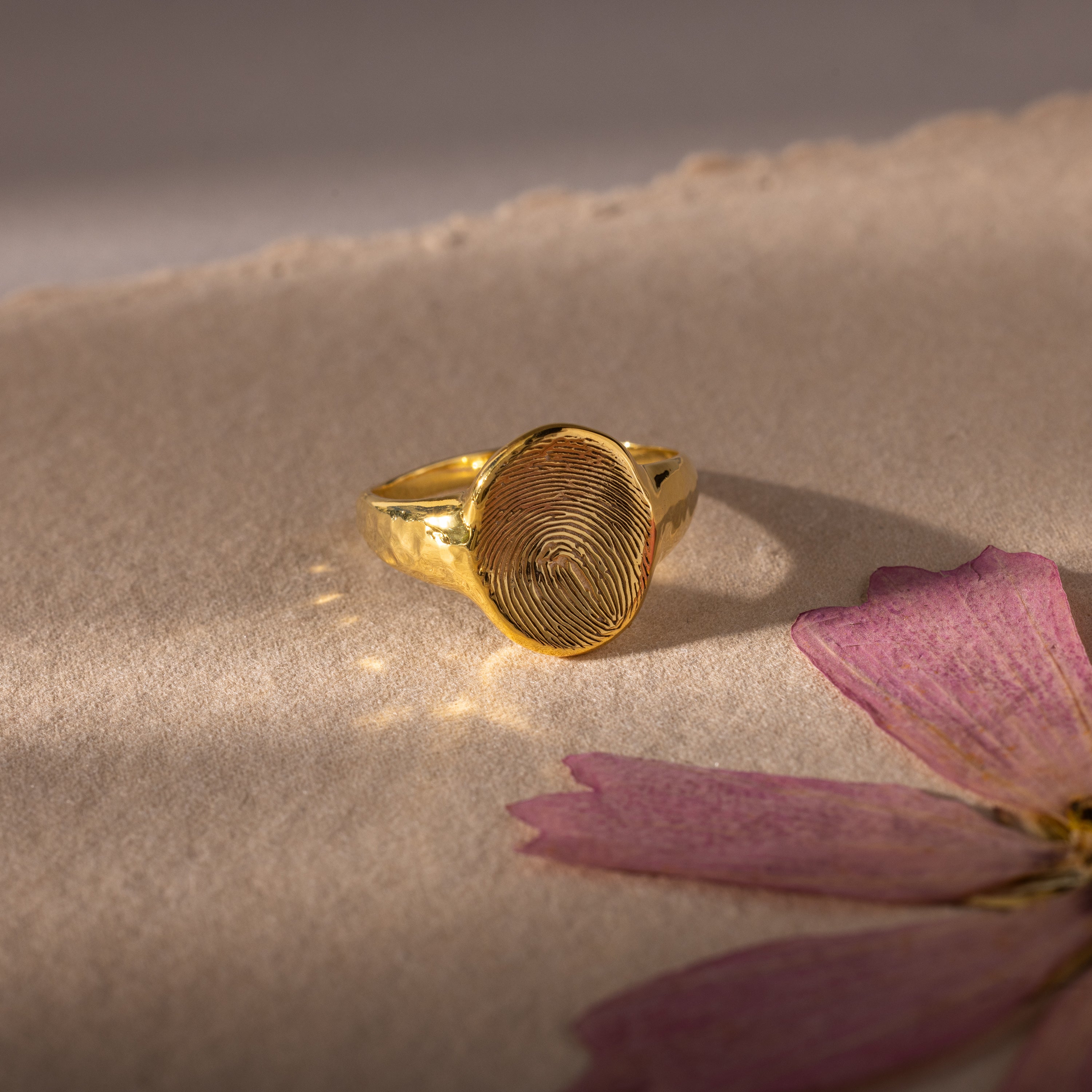 The Organic Fingerprint Signet Ring in 18K Gold rests on textured paper beside a pink dried flower, creating a stunning personalized jewelry keepsake.