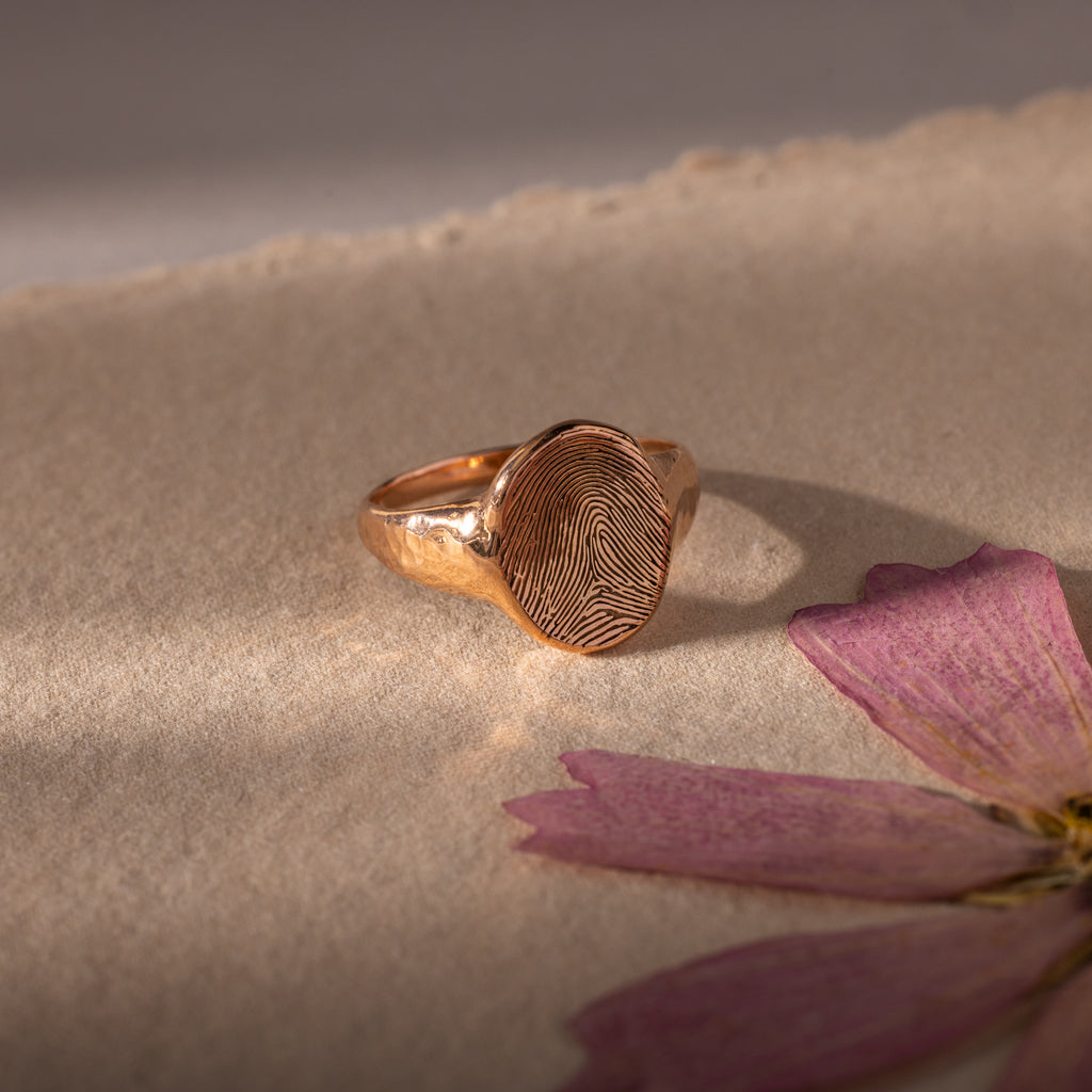 The Organic Fingerprint Signet Ring in Rose Gold features a unique fingerprint design on a textured surface, creating an elegant, personalized keepsake. Shown alongside dried pink flower petals, it beautifully captures special memories.