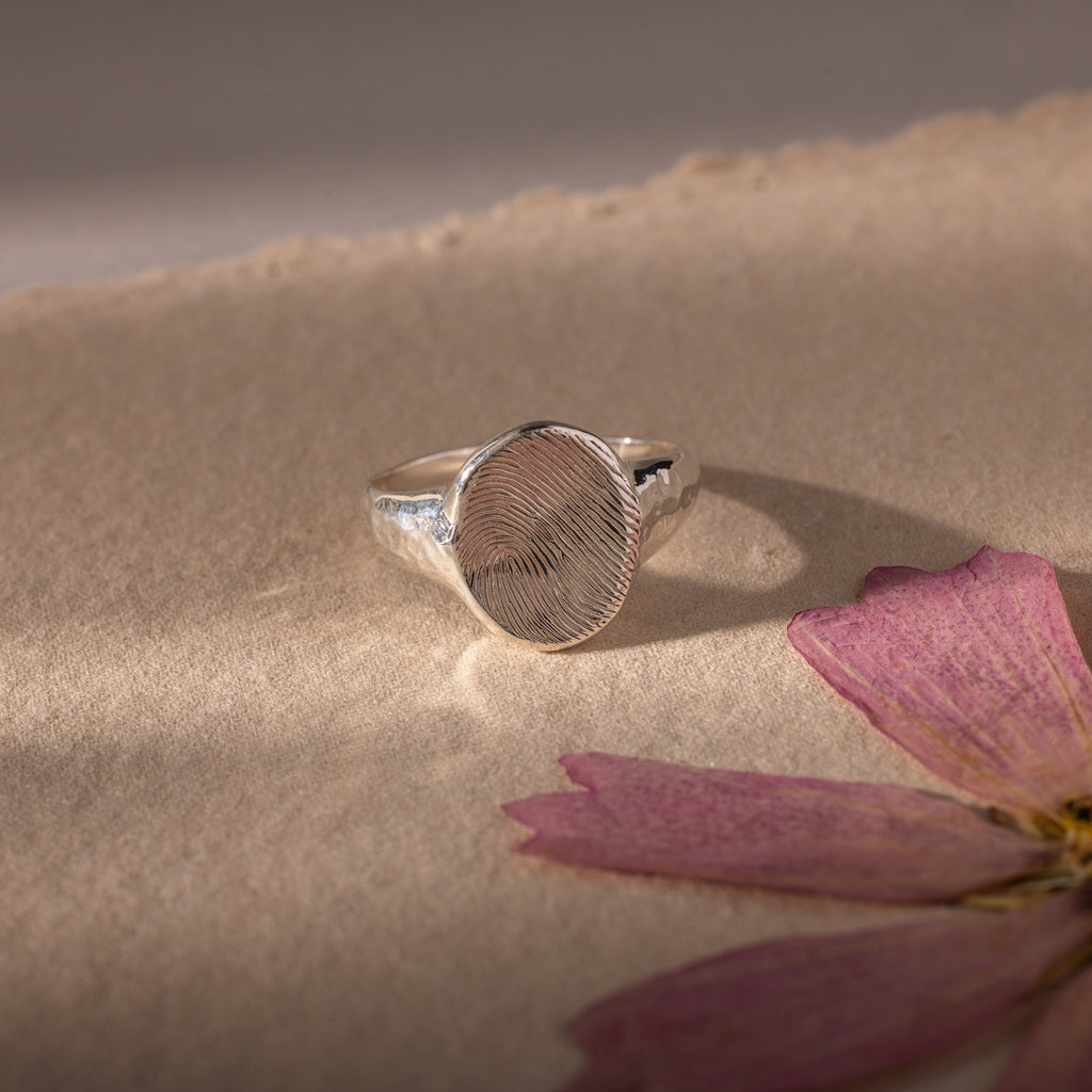 The Organic Fingerprint Signet Ring in Sterling Silver, featuring a unique design, rests on textured paper beside a dried pink flower petal—a beautiful, personalized jewelry piece and timeless keepsake.