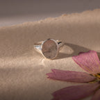 The Organic Fingerprint Signet Ring in Sterling Silver, featuring a unique design, rests on textured paper beside a dried pink flower petal—a beautiful, personalized jewelry piece and timeless keepsake.