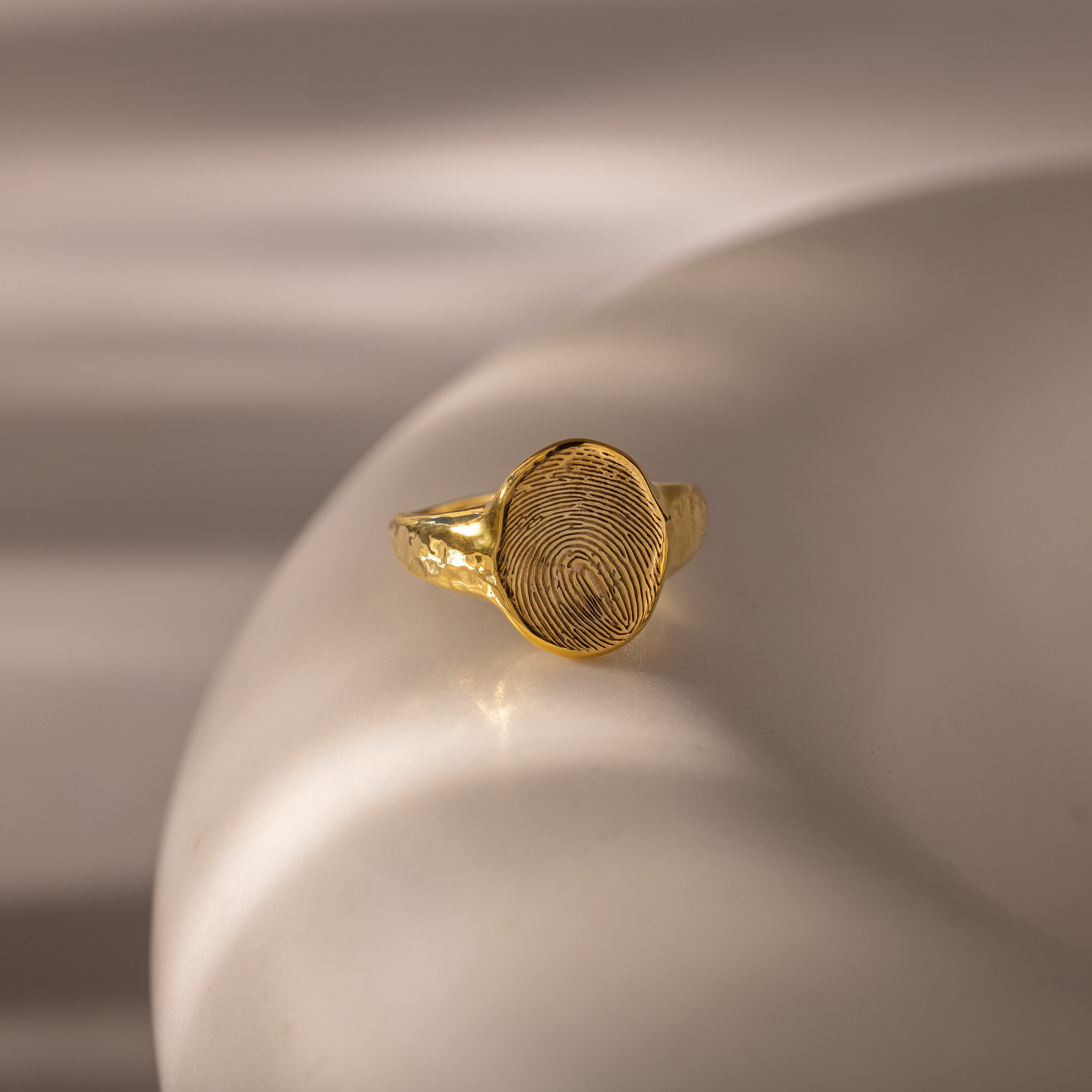 The Organic Fingerprint Signet Ring in 18K Gold is a personalized jewelry piece, showcased on a smooth, ivory-colored surface.