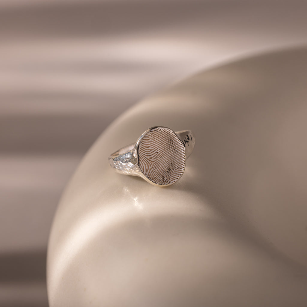 Organic Fingerprint Signet Ring: This handcrafted silver ring features a unique fingerprint design, displayed on a smooth beige surface with soft lighting for an elegant, personalized keepsake.