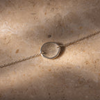A Dainty Organic Fingerprint Bracelet in Sterling Silver with a circular charm engraved with a unique fingerprint, elegantly displayed on a beige stone surface—a refined piece of personalized jewelry.