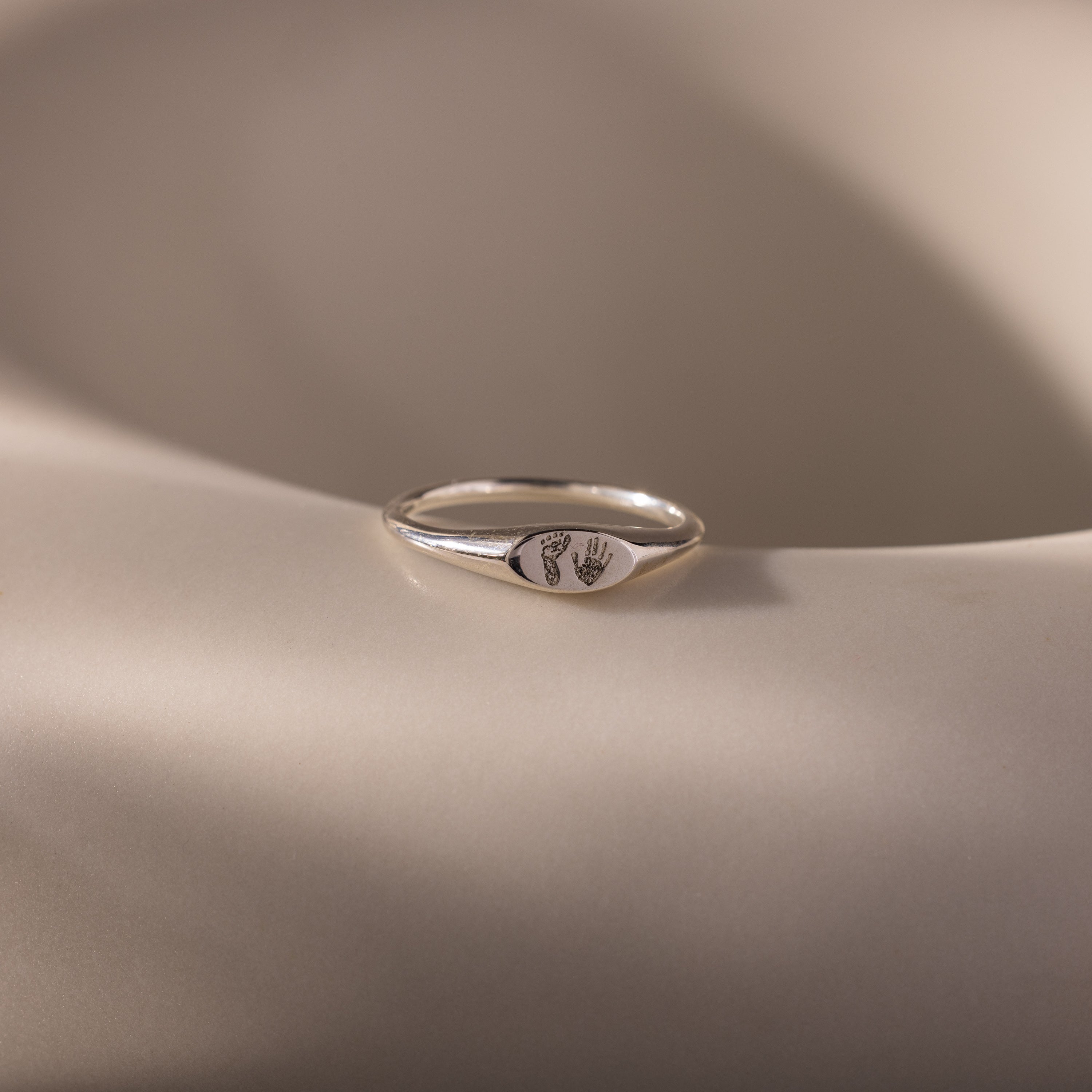 The Hand & Footprint Signet Ring in Sterling Silver, ideal as a baby keepsake, is displayed on a smooth beige surface.