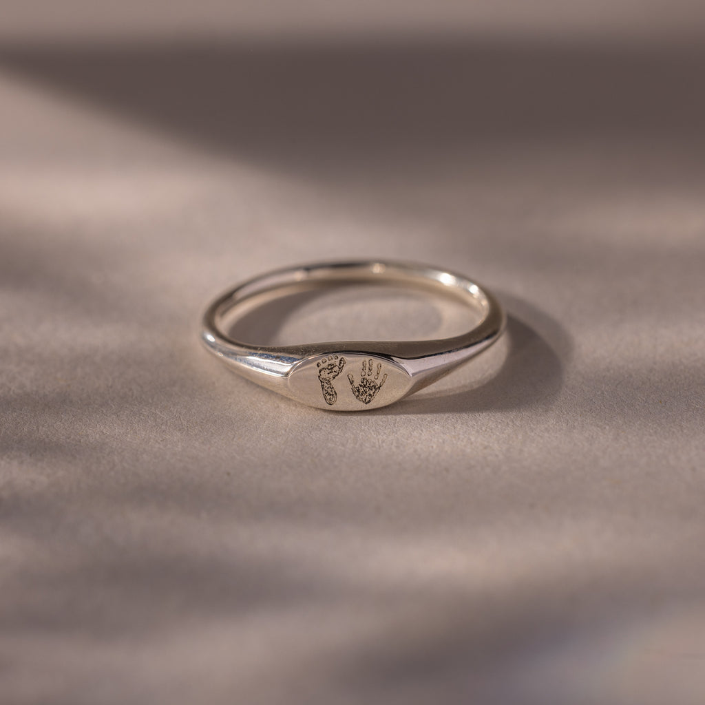 The Hand & Footprint Signet Ring in Sterling Silver features tiny engraved hand and footprints on a polished surface—ideal as a cherished baby keepsake, beautifully displayed on a light background.