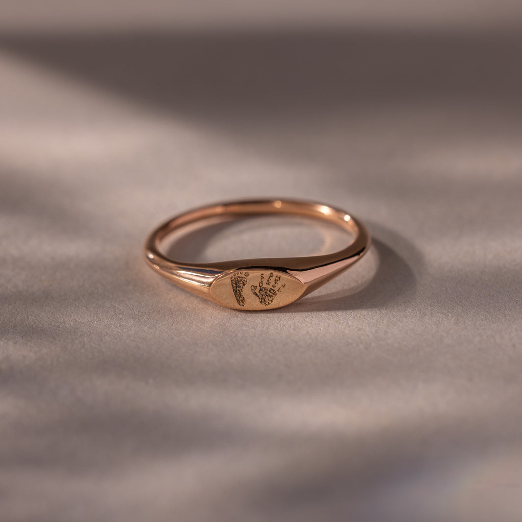 The Hand & Footprint Signet Ring in Rose Gold, engraved with tiny baby footprints, rests on a smooth, neutral surface—a perfect keepsake.