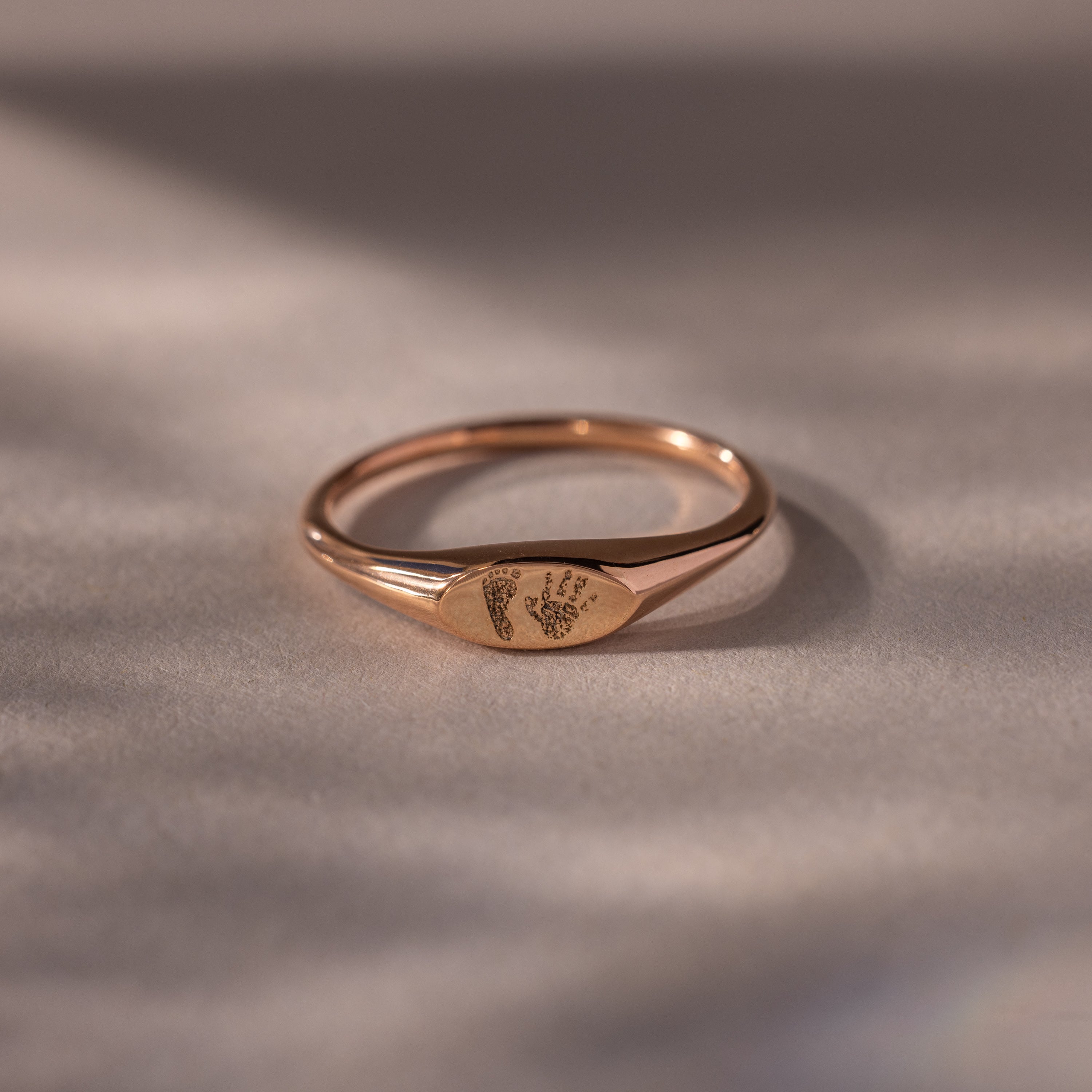 The Hand & Footprint Signet Ring in Rose Gold, engraved with tiny baby footprints, rests on a smooth, neutral surface—a perfect keepsake.