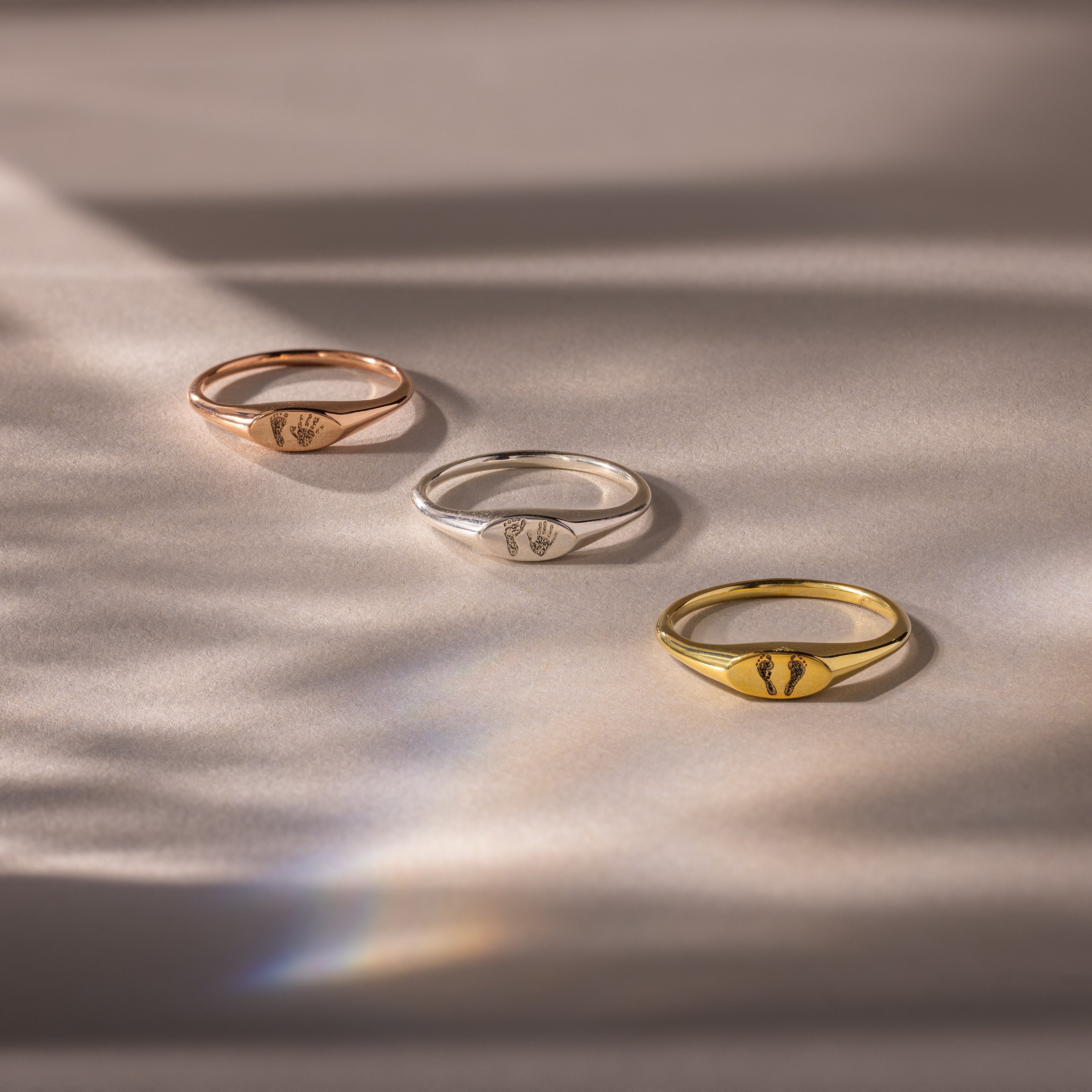 The Hand & Footprint Signet Ring in Rose Gold is shown among silver and gold rings, all arranged on a soft, light background with gentle shadows.