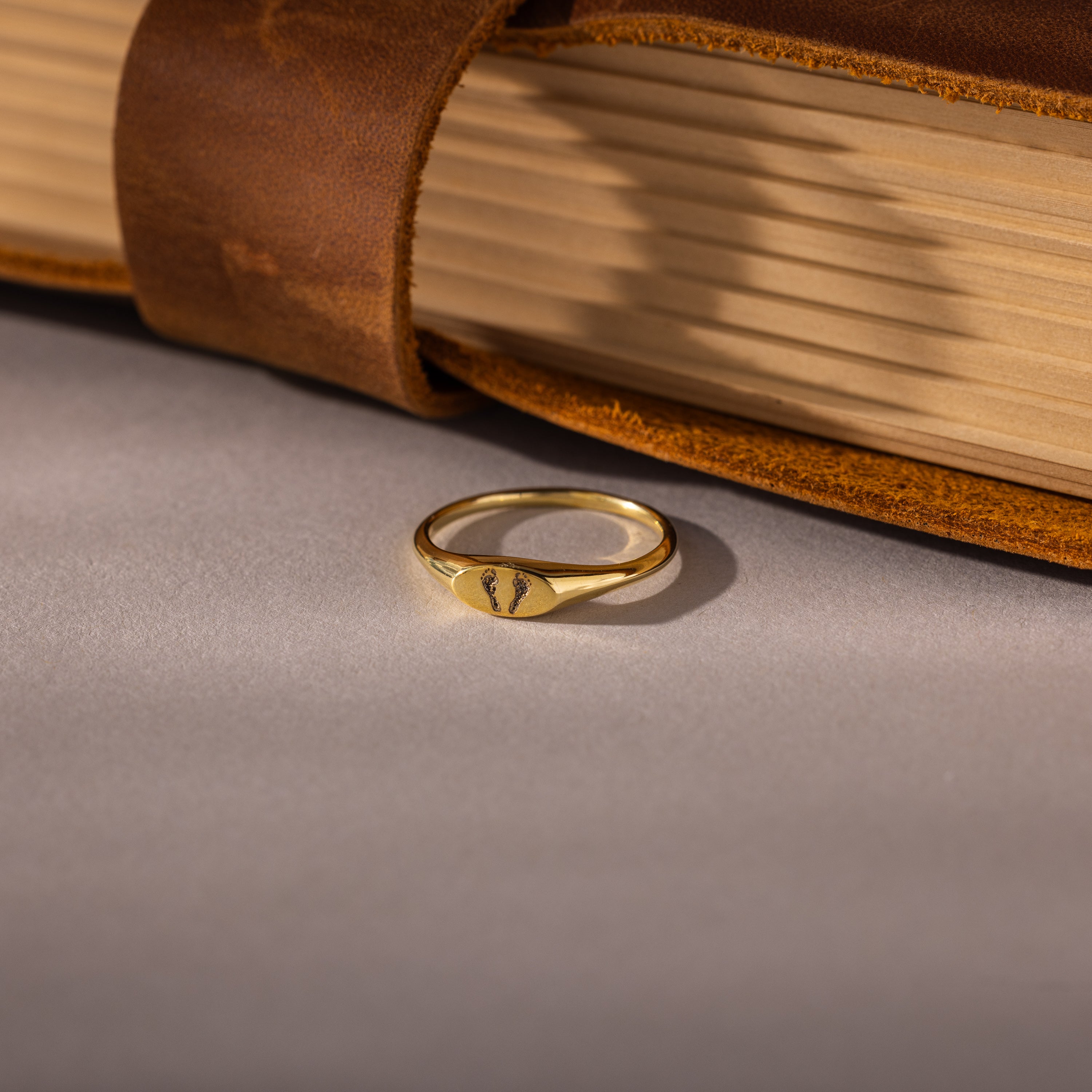 The Hand & Footprint Signet Ring, featuring an engraved design, rests beside a brown leather-covered closed book—an ideal piece for keepsake jewelry.