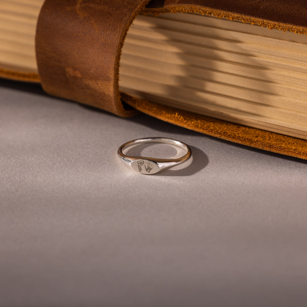 The Hand & Footprint Signet Ring in Sterling Silver sits on a gray surface beside a leather-bound book—a timeless baby keepsake to treasure special memories.