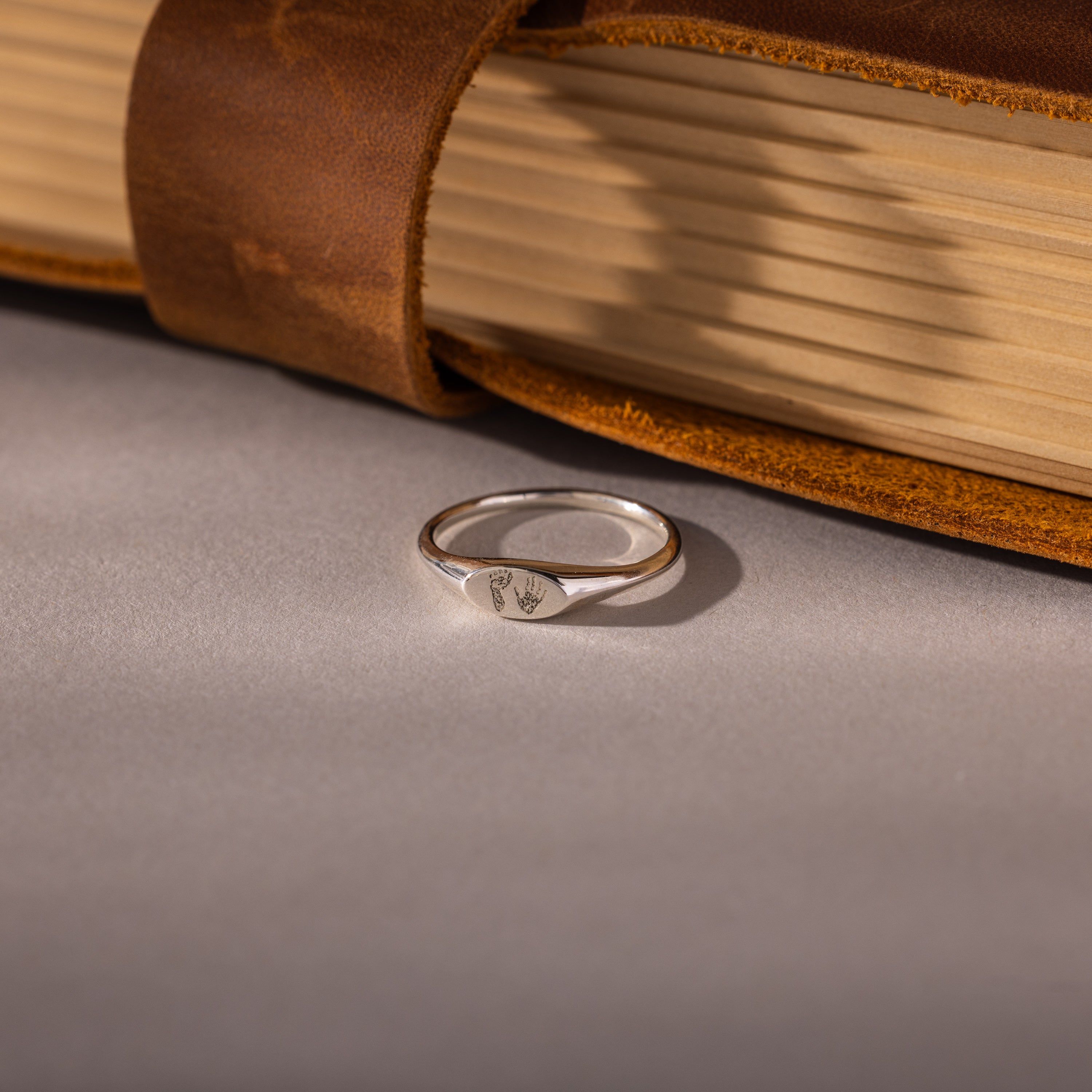The Hand & Footprint Signet Ring in Sterling Silver sits on a gray surface beside a leather-bound book—a timeless baby keepsake to treasure special memories.