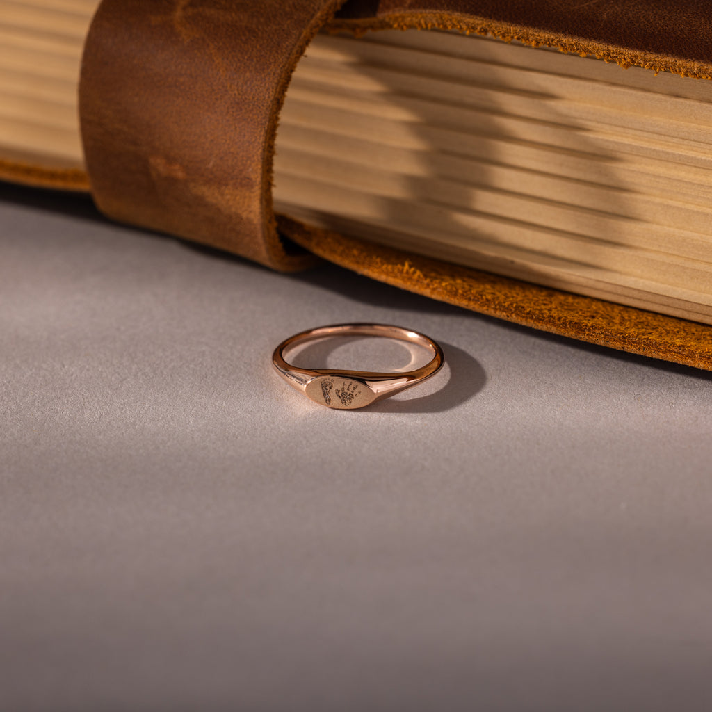 The Hand & Footprint Signet Ring in Rose Gold rests on a grey surface beside a closed brown leather-bound book, making a timeless baby keepsake.