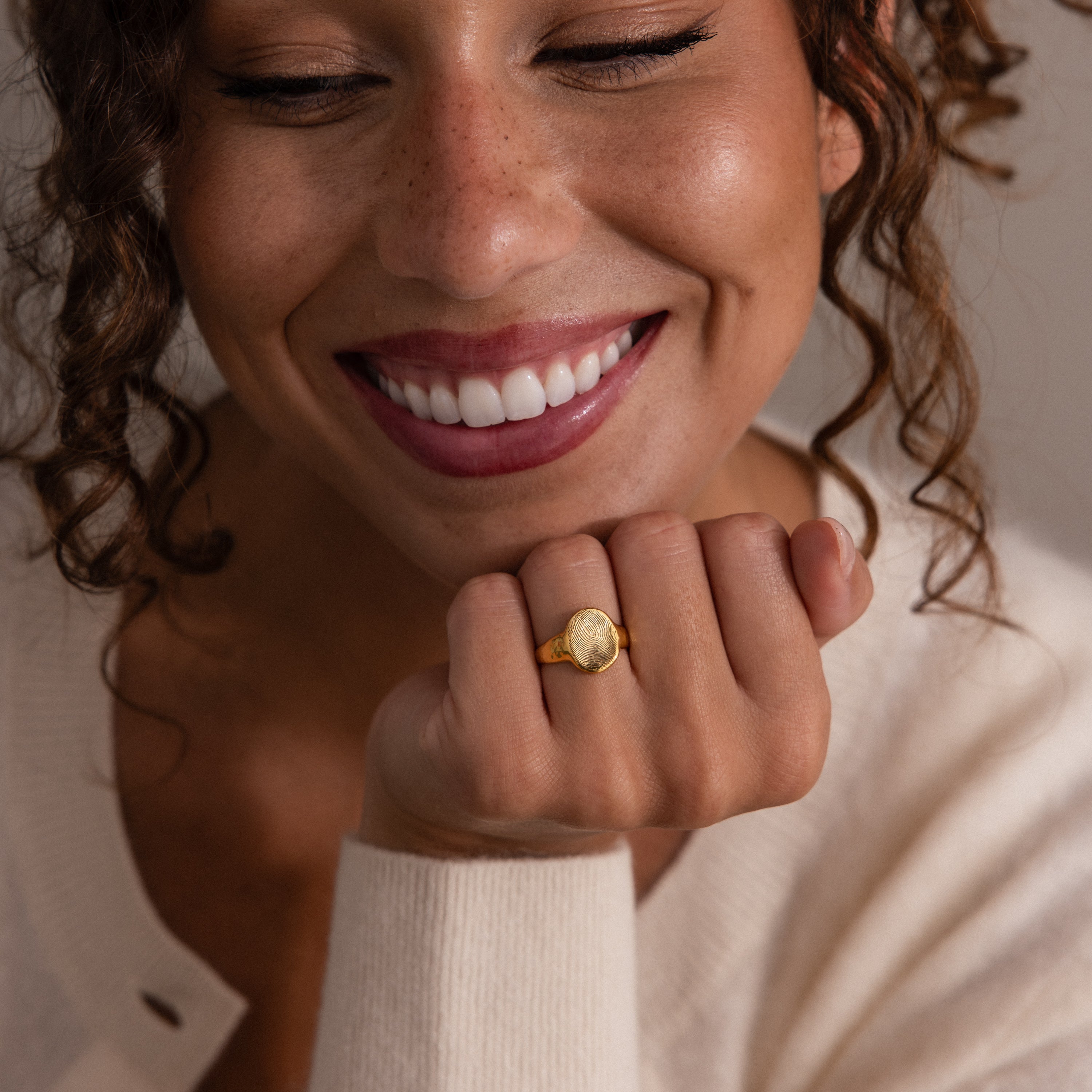 A smiling woman with curly hair showcases the Organic Fingerprint Signet Ring in Rose Gold on her hand, paired with a cream sweater—a stunning, personalized fingerprint jewelry piece and a timeless keepsake.