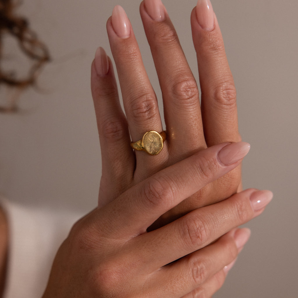 Two hands with manicured nails, one showcasing the Organic Fingerprint Signet Ring in 18K Gold, are held gracefully against a neutral background.