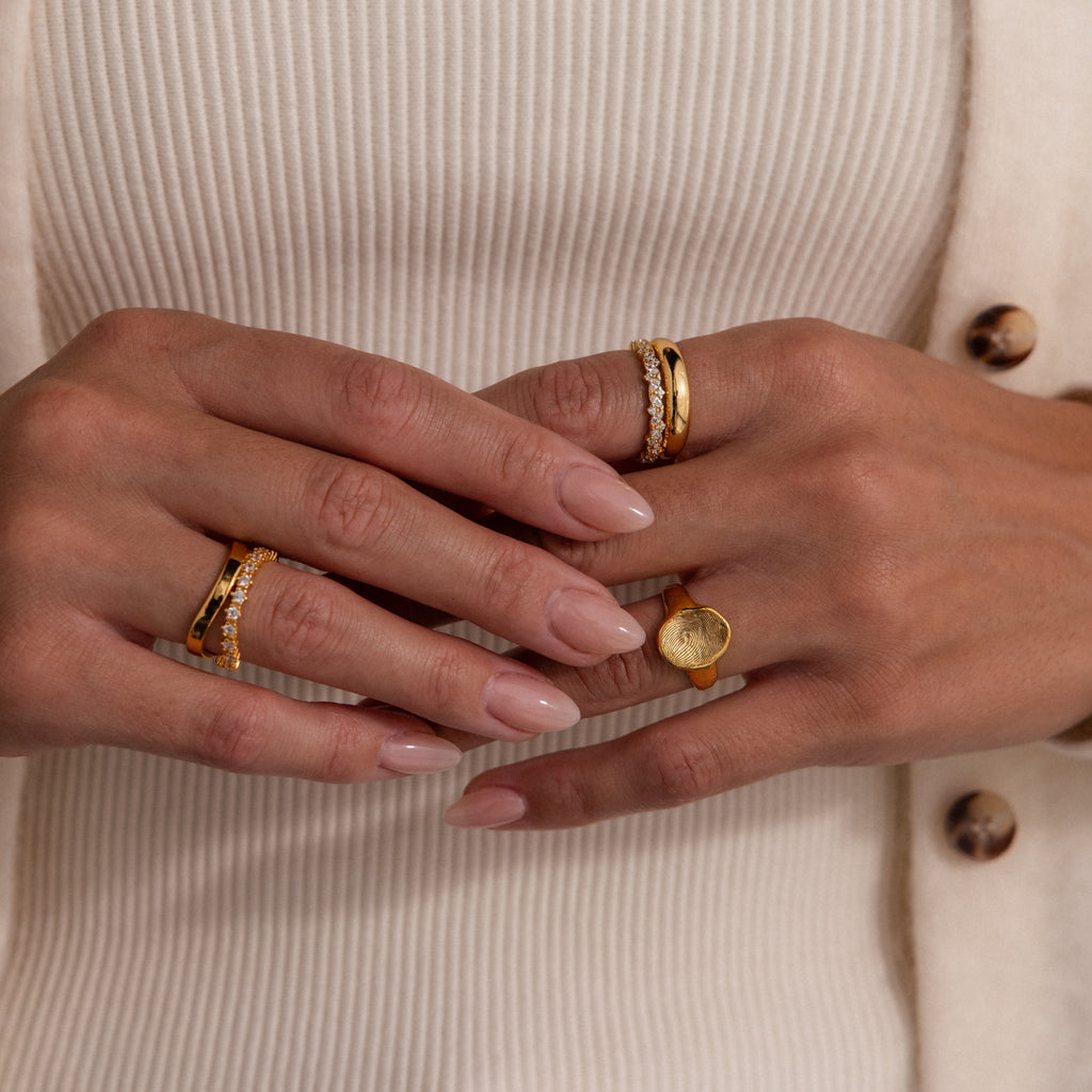 A woman's hands with nude nails wearing the Organic Fingerprint Signet Ring in 18K Gold and diamond bands, shown against a cream ribbed top and cardigan.