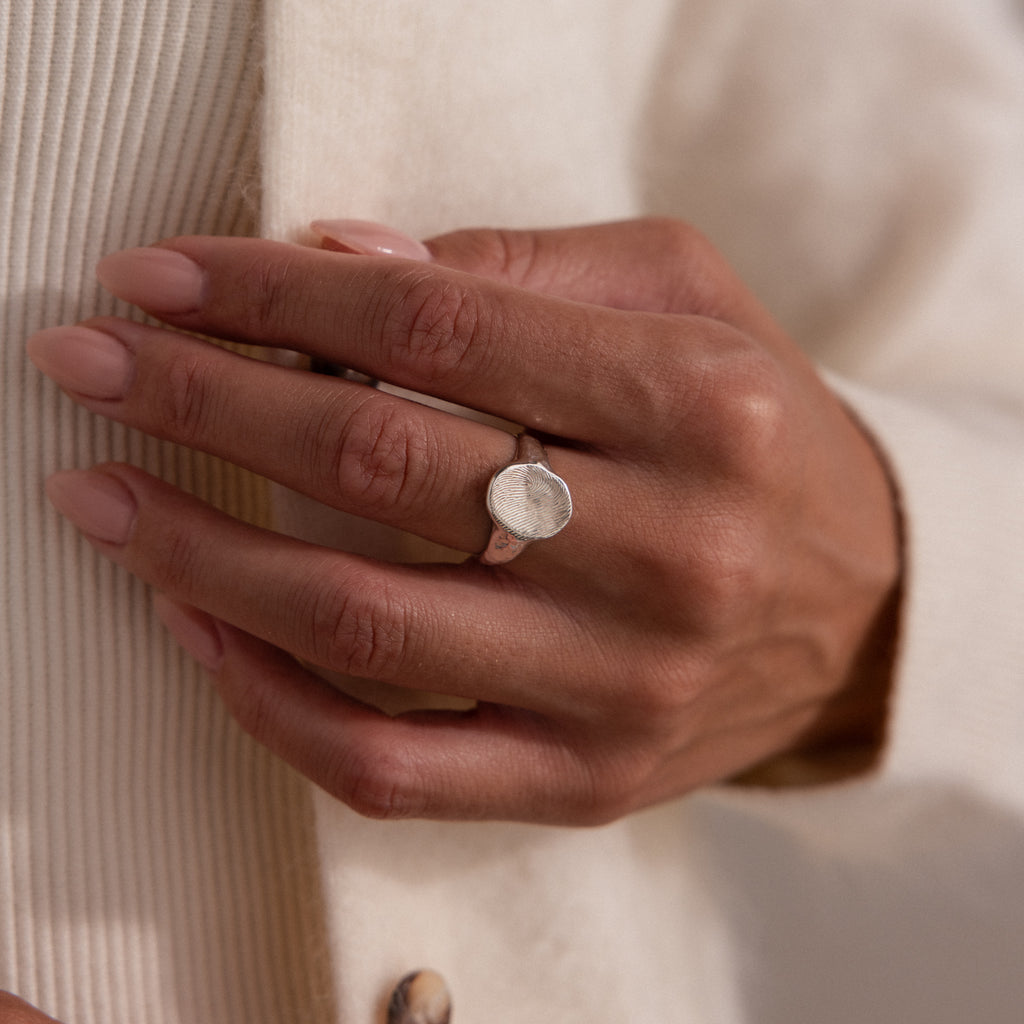 A hand with manicured nails wears the Organic Fingerprint Signet Ring in Sterling Silver, resting on a cream-toned textured garment—an elegant, personalized jewelry piece.