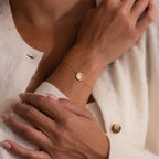 A woman in a cream-colored sweater wears the Dainty Organic Fingerprint Bracelet in Rose Gold, featuring a round pendant.