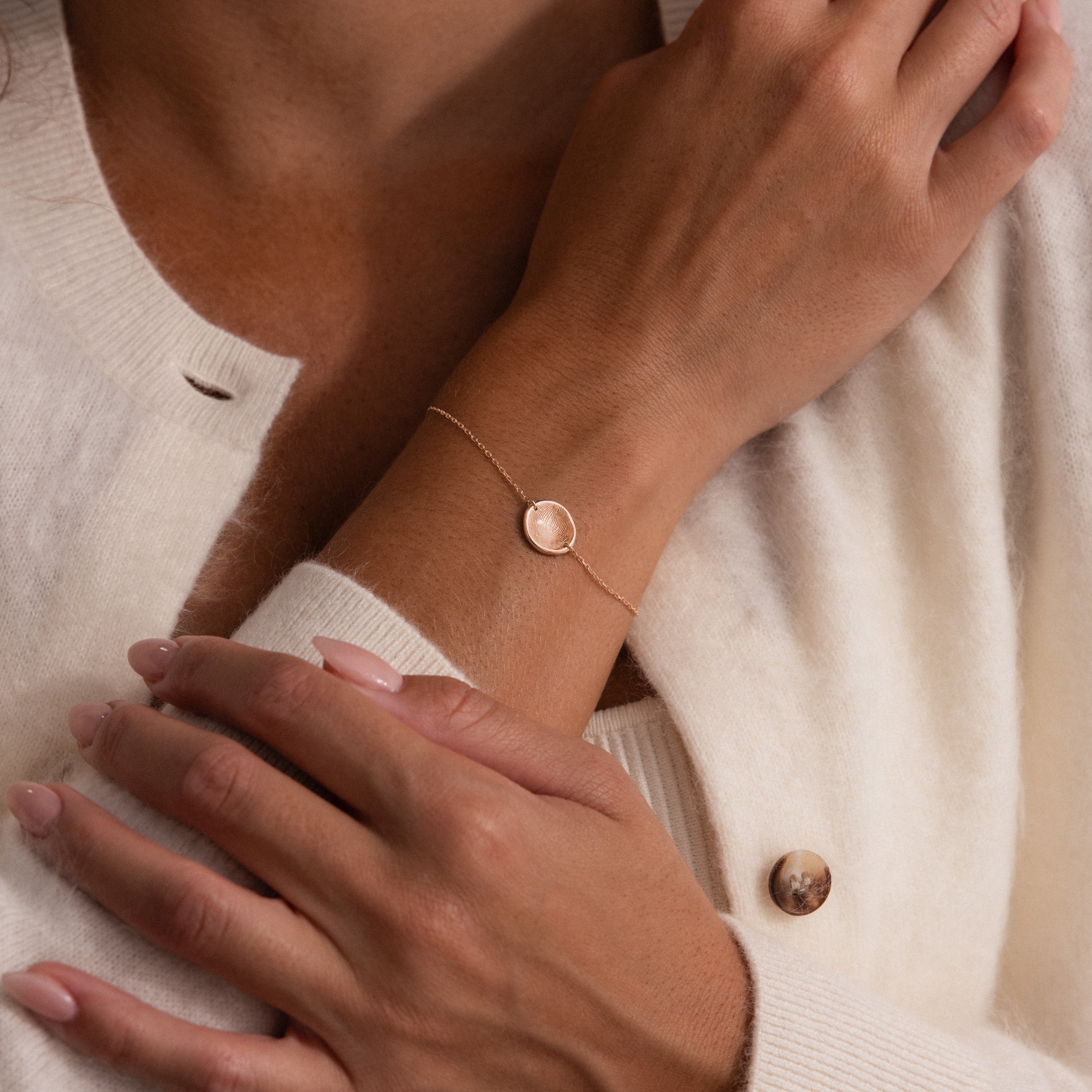 A woman in a cream-colored sweater wears the Dainty Organic Fingerprint Bracelet in Rose Gold, featuring a round pendant.