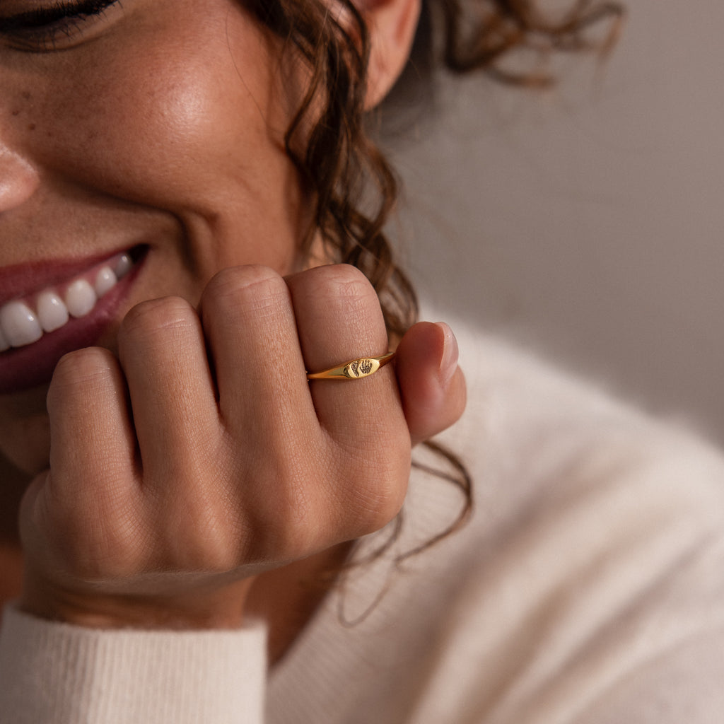 A smiling woman wears the Hand & Footprint Signet Ring in 18K Gold, engraved with a unique design, and rests her hand against her face.
