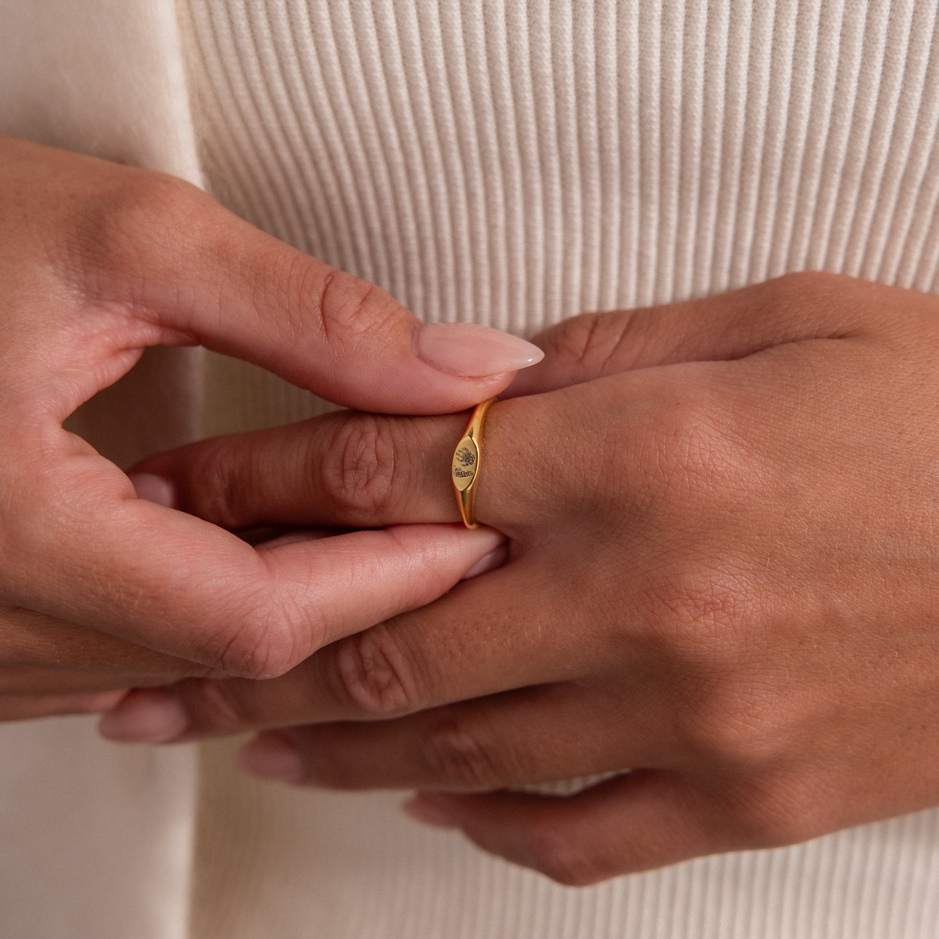 A person with manicured nails wears the Hand & Footprint Signet Ring in 18K Gold on their finger, set against a beige ribbed fabric background.
