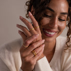 A smiling woman with curly hair displays gold rings, including the Hand & Footprint Signet Ring, on her fingers while wearing a cream sweater.