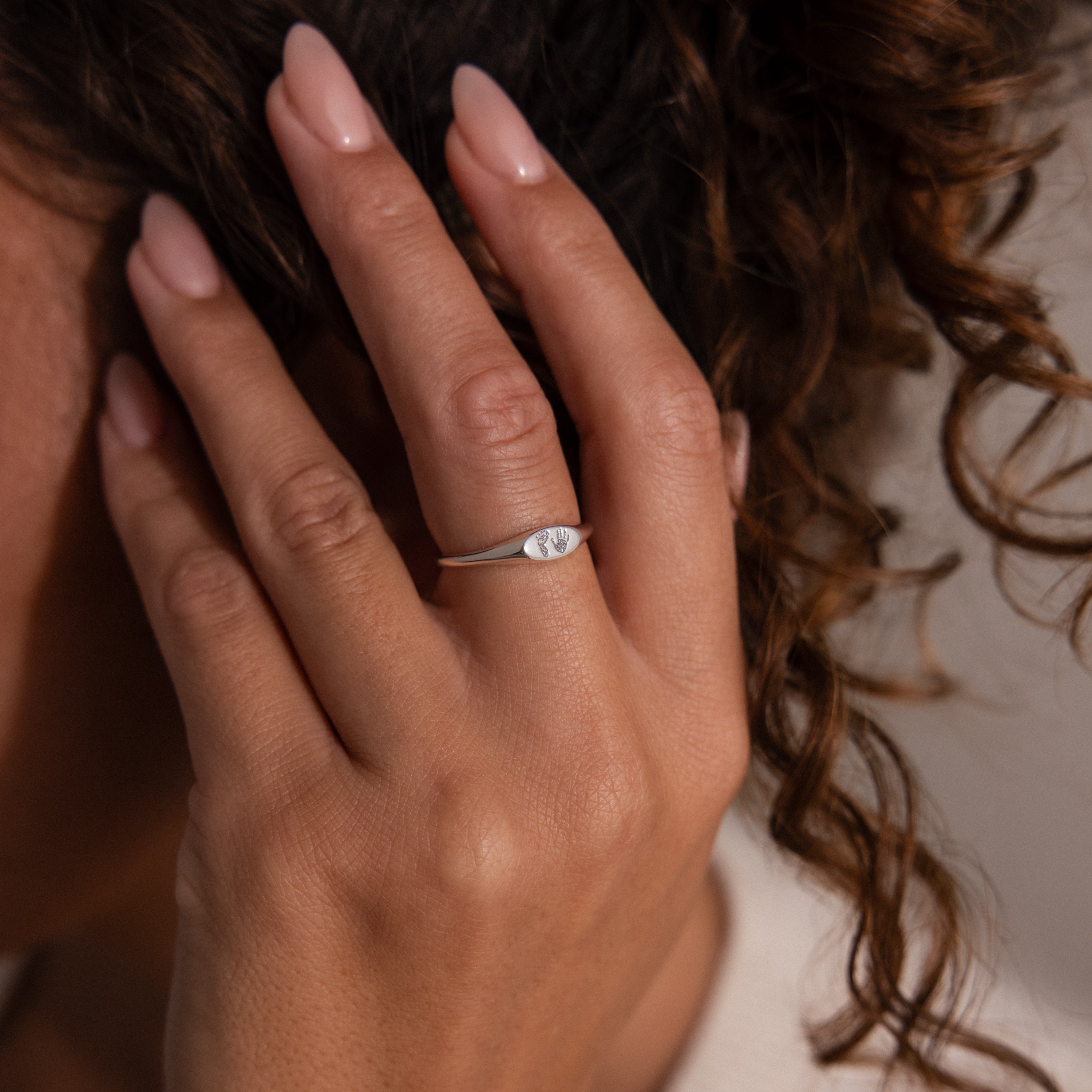 A hand with manicured nails wears the Hand & Footprint Signet Ring in Sterling Silver next to curly brown hair.